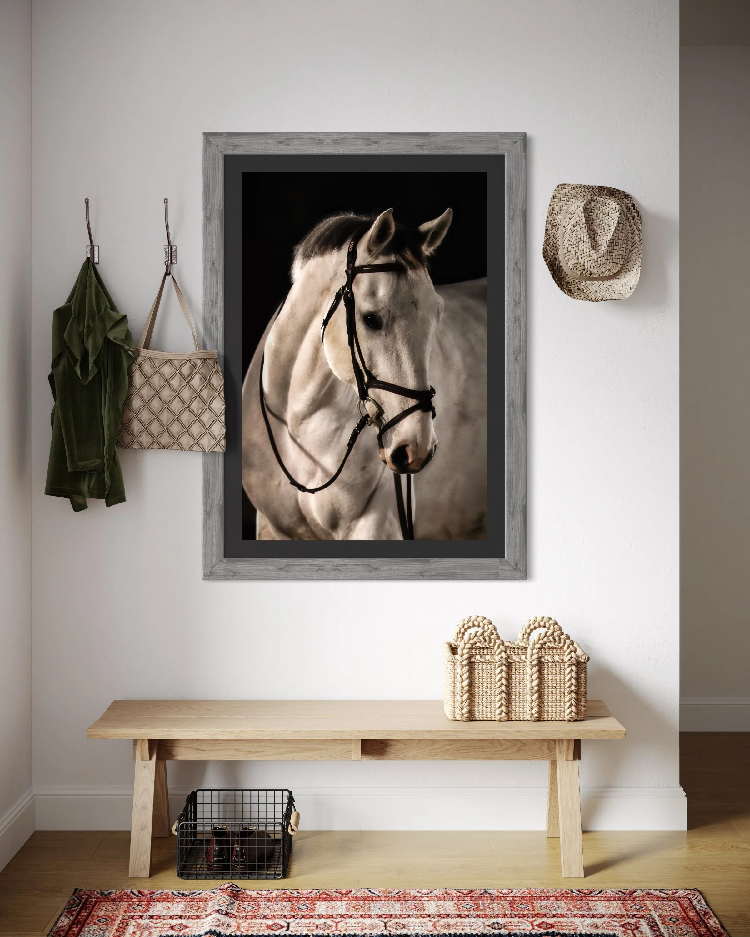 Interior wall with a large framed black and white photograph of a white horse, a straw hat hanging on the wall, a green shirt and quilted purse hanging on hooks, a wooden bench with woven baskets on top, and a small wire basket with shoes underneath, on a patterned rug.