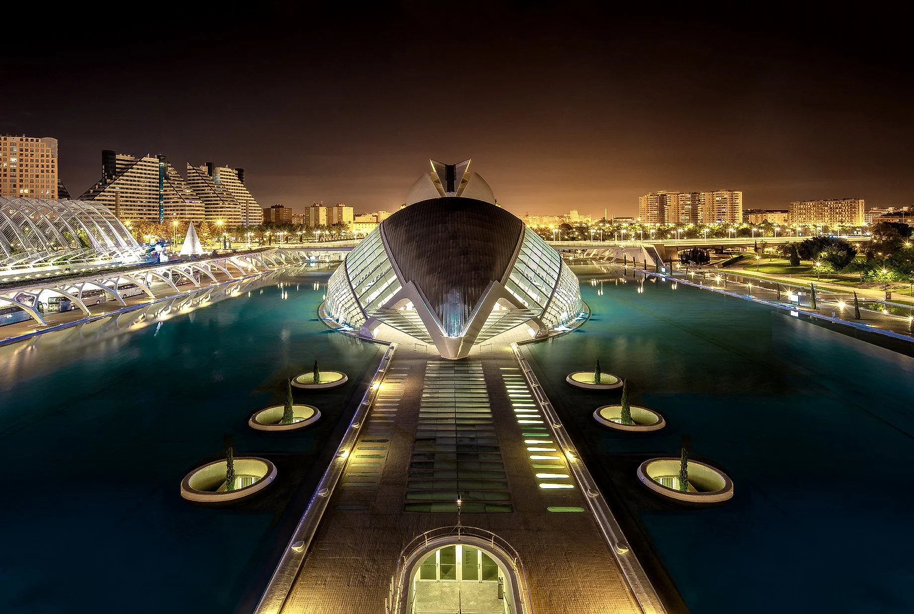 Night view of a modern, illuminated building resembling a whale, surrounded by water with city buildings in the background.