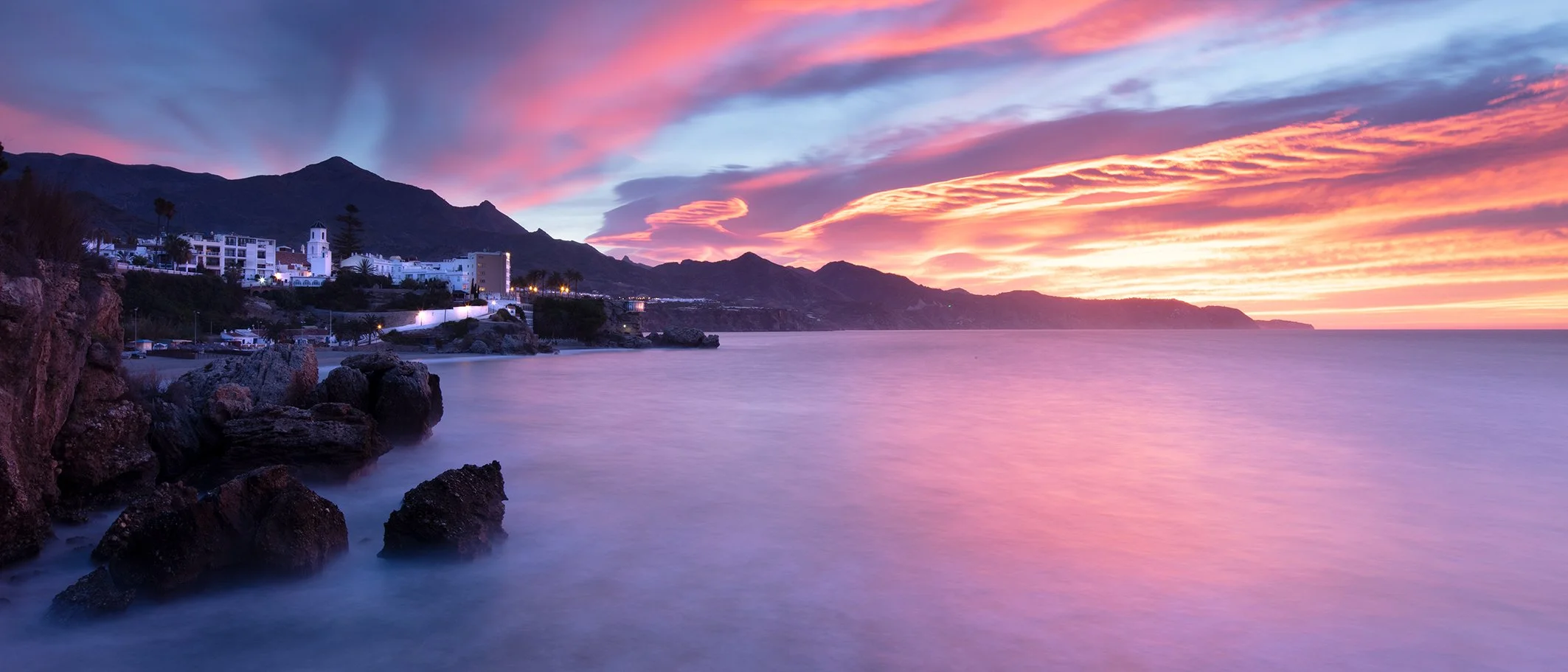 Sunset over a coastal town with purple and pink clouds, rocky shoreline, calm water, and mountains in the background.