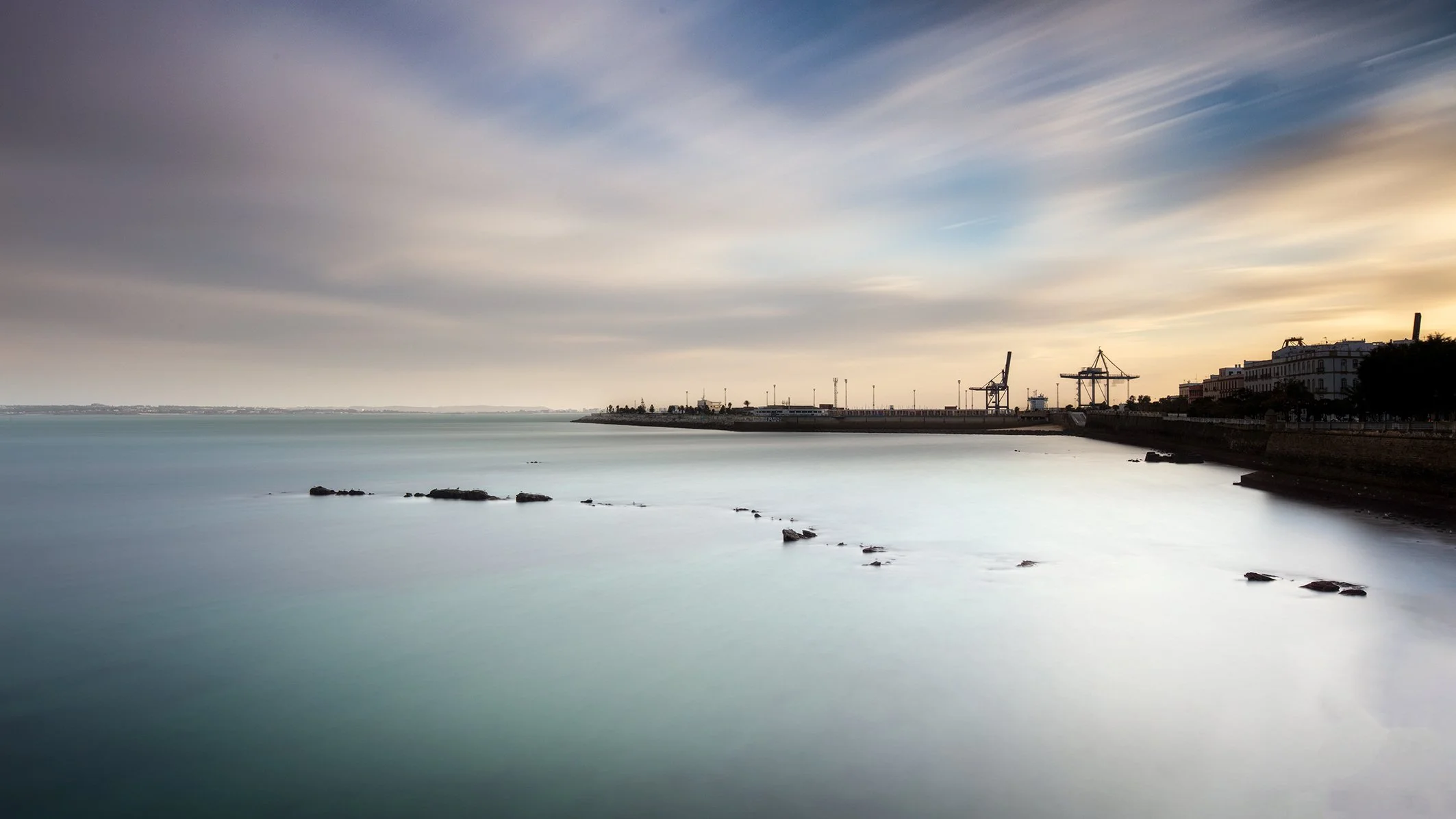 A calm harbor scene during sunset with a long exposure effect, showing the water surface, some rocks, a distant city shoreline, industrial cranes, and cloudy sky.