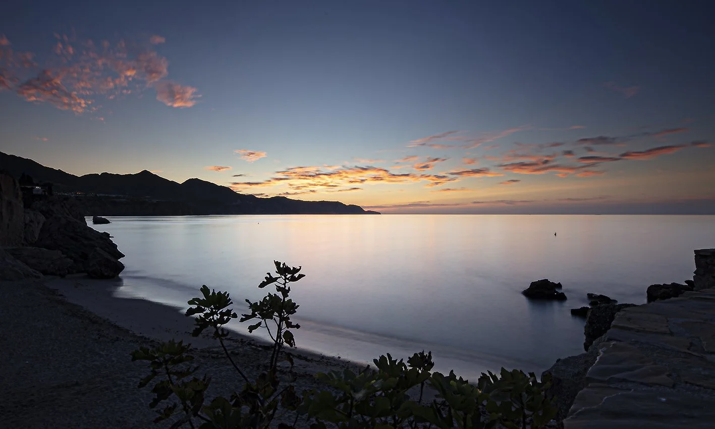 Sunset over calm ocean with a mountain silhouette in the distance and rocks along the shoreline