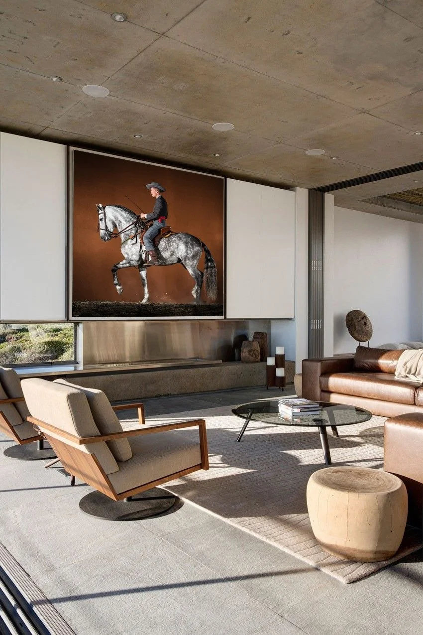 Modern living room with a large portrait of a rider on a gray horse on the wall, a brown leather sofa, beige armchairs, a glass coffee table, and a wooden stool, with sunlight streaming in.