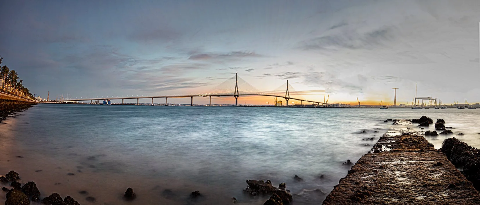 Long exposure photo of a bridge over a calm body of water during sunset with a partly cloudy sky and industrial structures in the distance.
