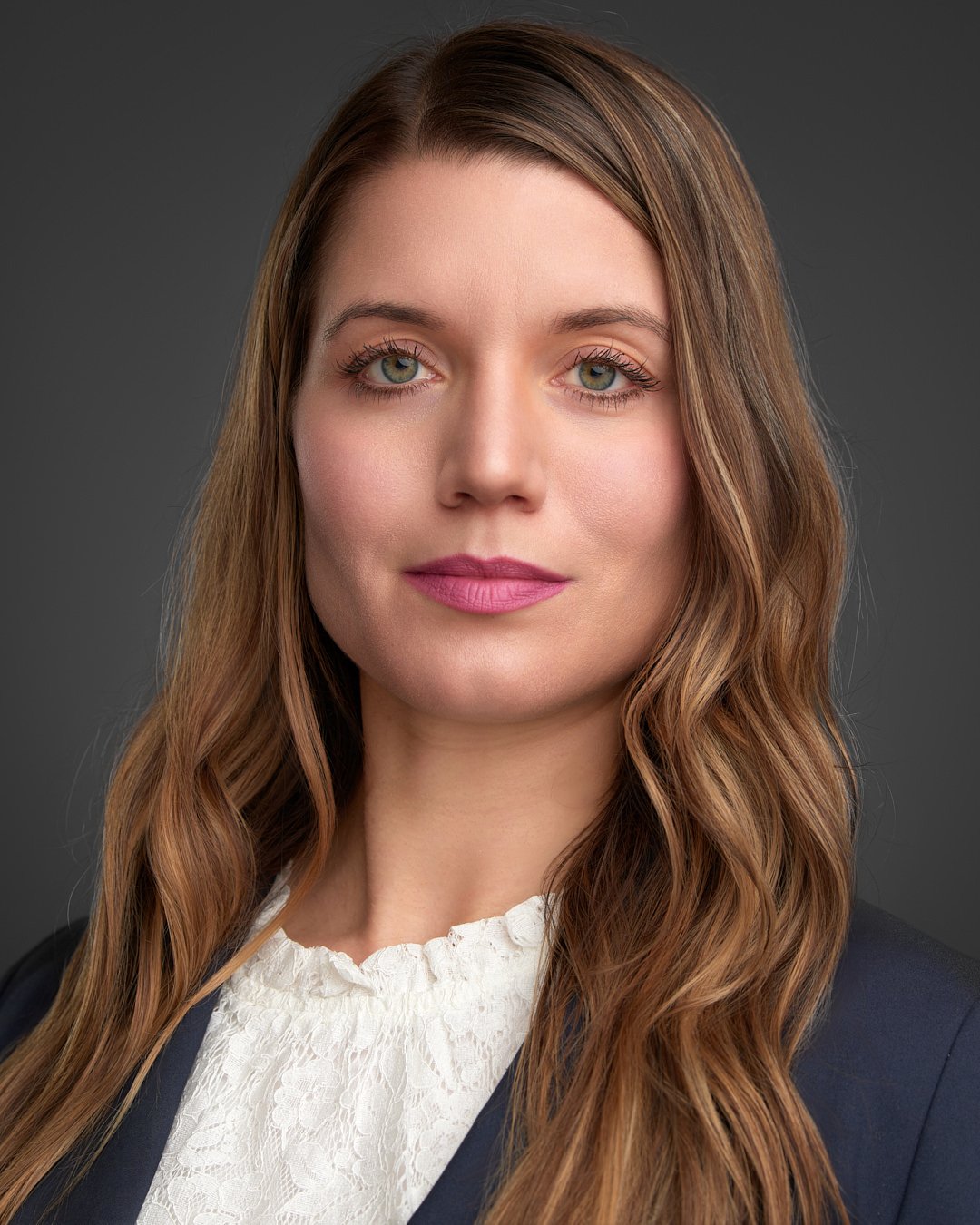 Polished headshot of a young woman with light wavy hair, wearing a white blouse against a dark gray backdrop