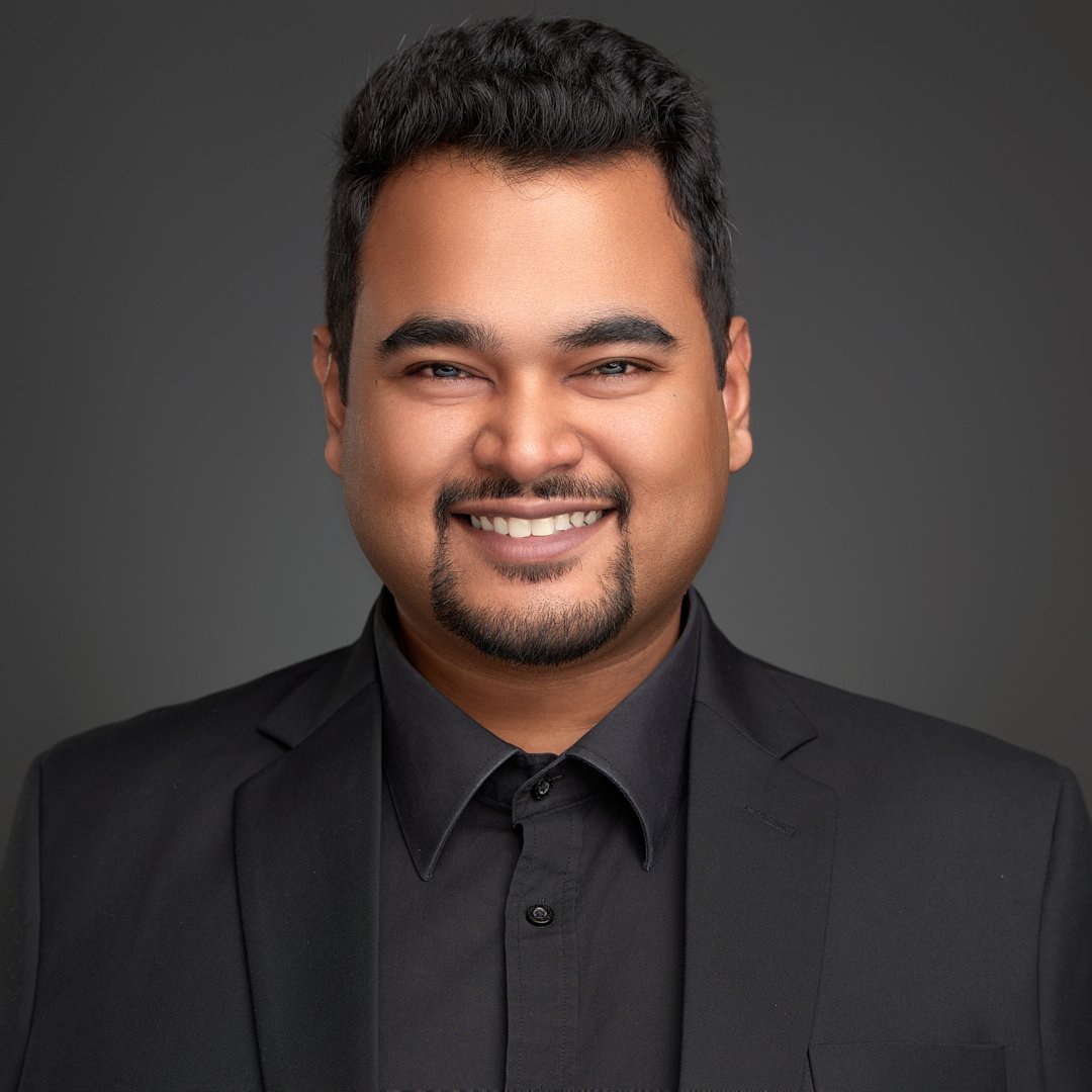 Clean corporate portrait of a South Asian man with a warm smile, wearing an all-black shirt and blazer