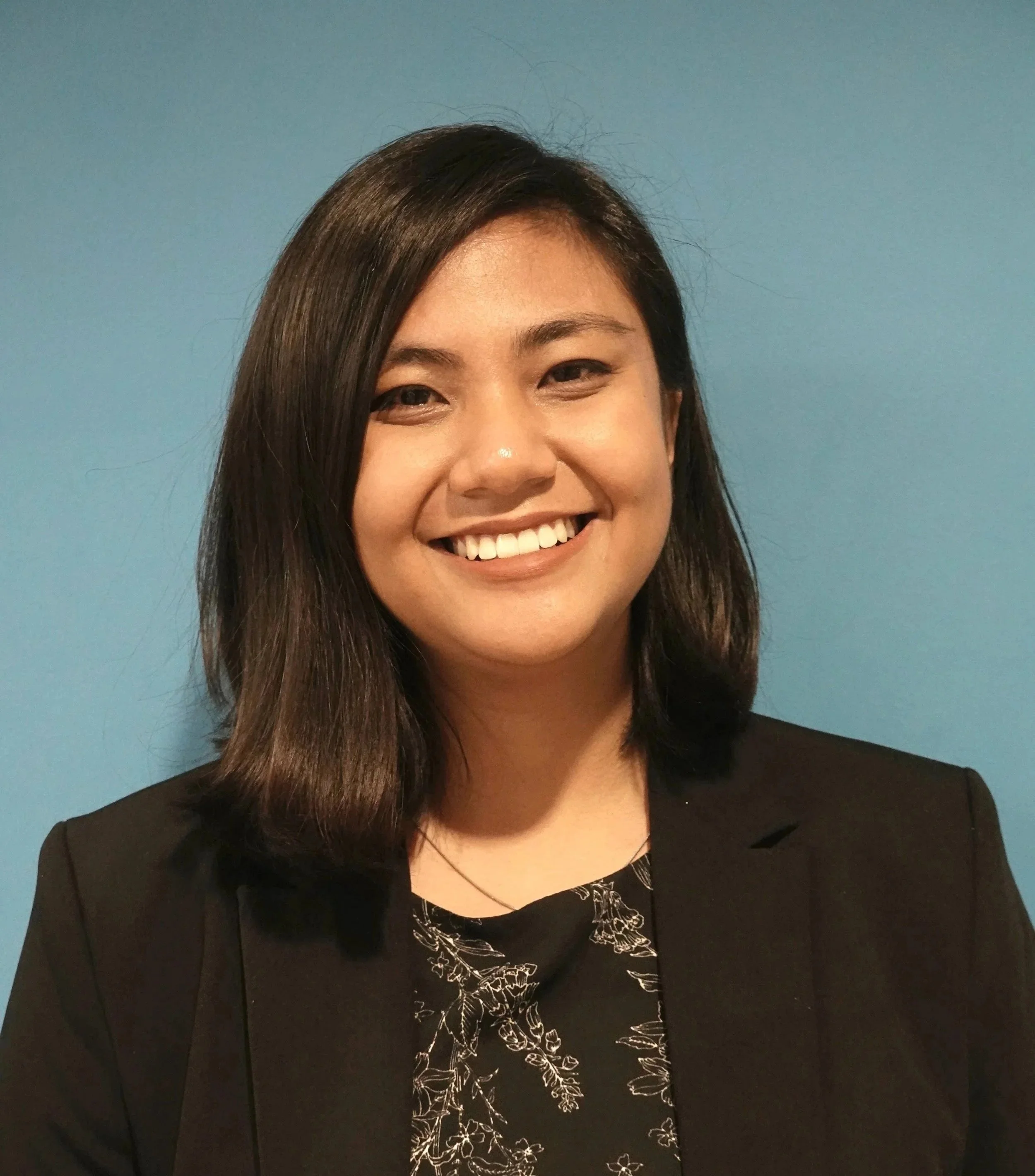 A young woman with straight black hair sitting on outdoor stairs next to a green metal railing, wearing a black jacket and jeans, smiling at the camera.
