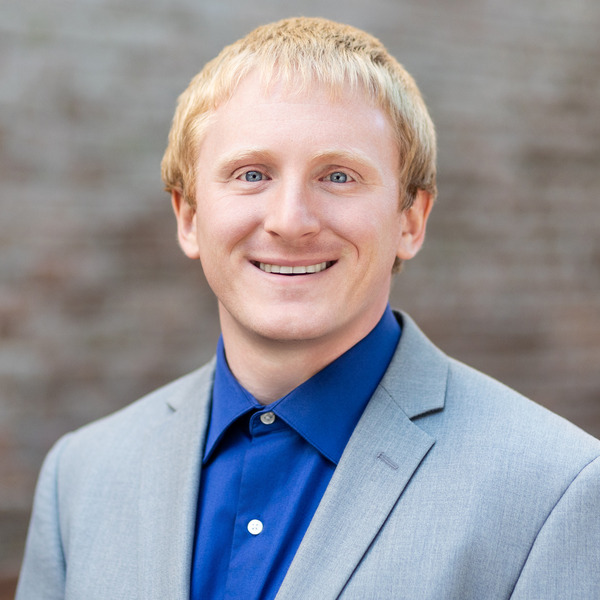A young man with red hair and blue eyes smiling, wearing a black shirt, with a blurred modern office background.