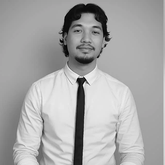 Black and white portrait of a young man with curly hair, a goatee, wearing a white dress shirt and a dark tie, smiling slightly, against a plain background.