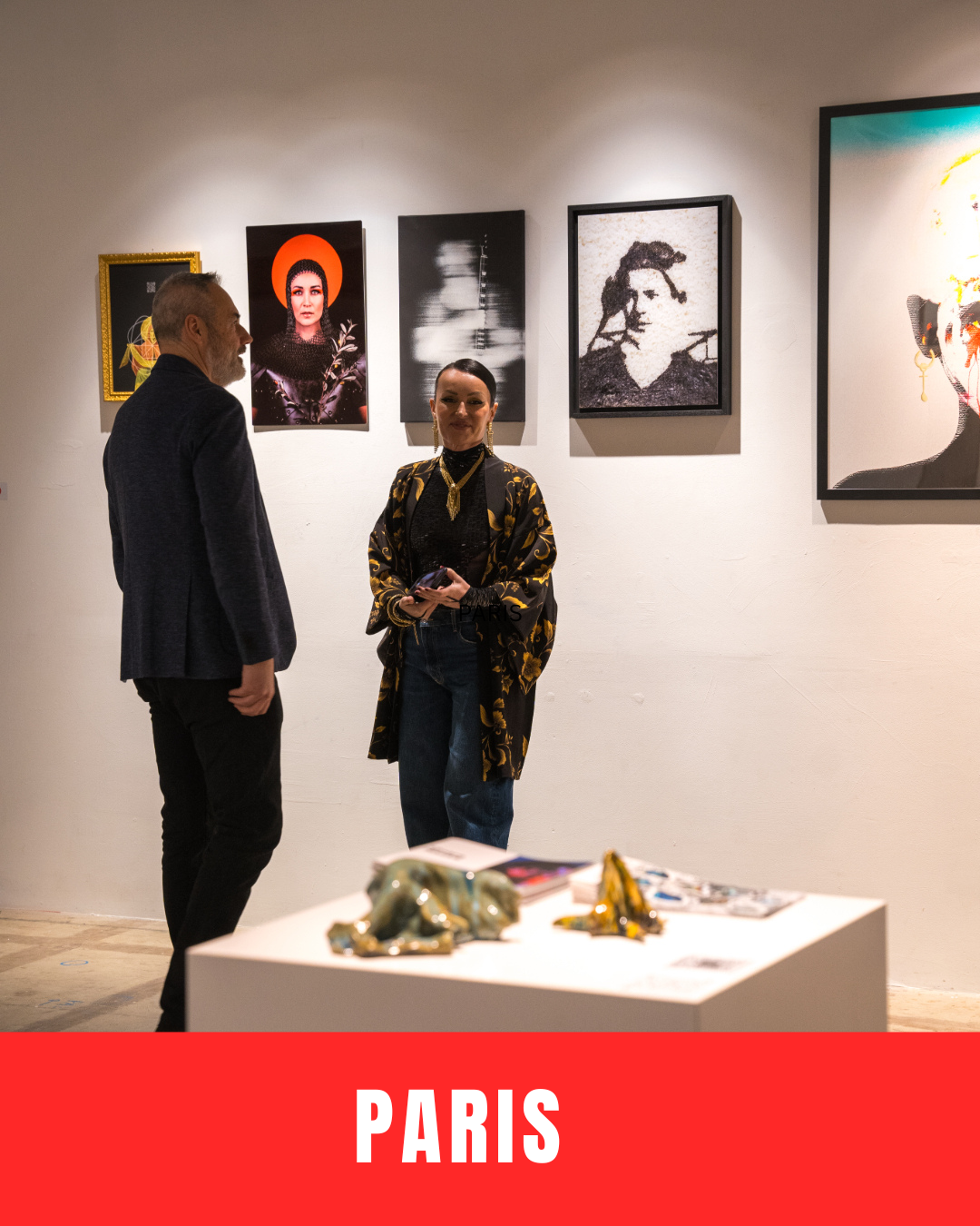 Two people are standing in an art gallery, engaging in conversation. The gallery wall behind them features various framed artworks, including portraits and abstract images. A white display table with sculptures and magazines is in the foreground. The word 'PARIS' appears at the bottom of the image.