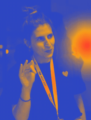 A woman with hair in a bun making an 'OK' sign with her hand, wearing a lanyard labeled 'The Holy Art' around her neck.