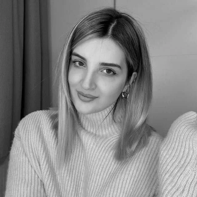 A young woman with shoulder-length hair winks at the camera, wearing a light-colored turtleneck sweater and hoop earrings, in a black and white photo.