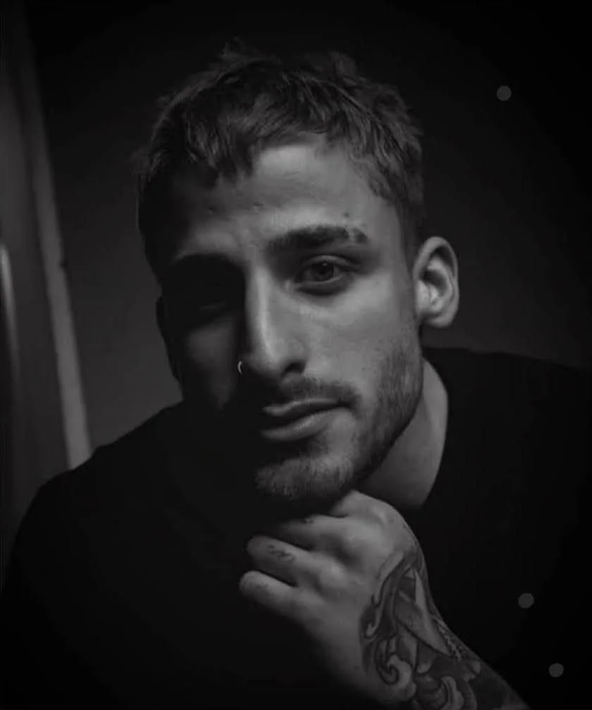 Black and white close-up portrait of a young man with short hair, light facial hair, and a nose piercing, resting his chin on his hand which has visible tattoos.