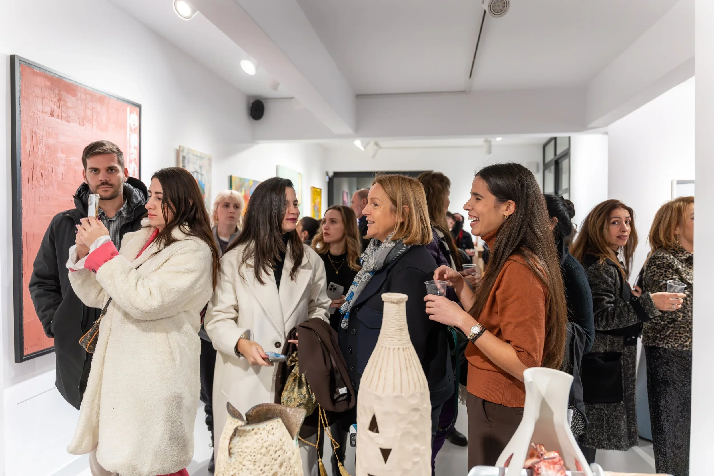 People viewing artwork at an art gallery, engaged in conversation, some holding drinks, with various art pieces on the walls and ceramic sculptures on display in the foreground.