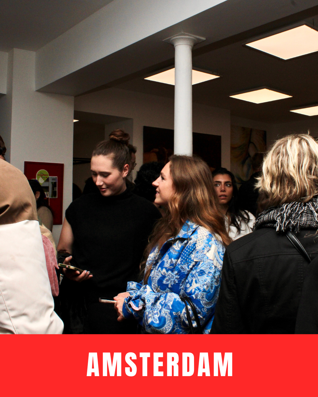 Group of people socializing at an indoor event with modern ceiling lights and a white pillar, in Amsterdam.