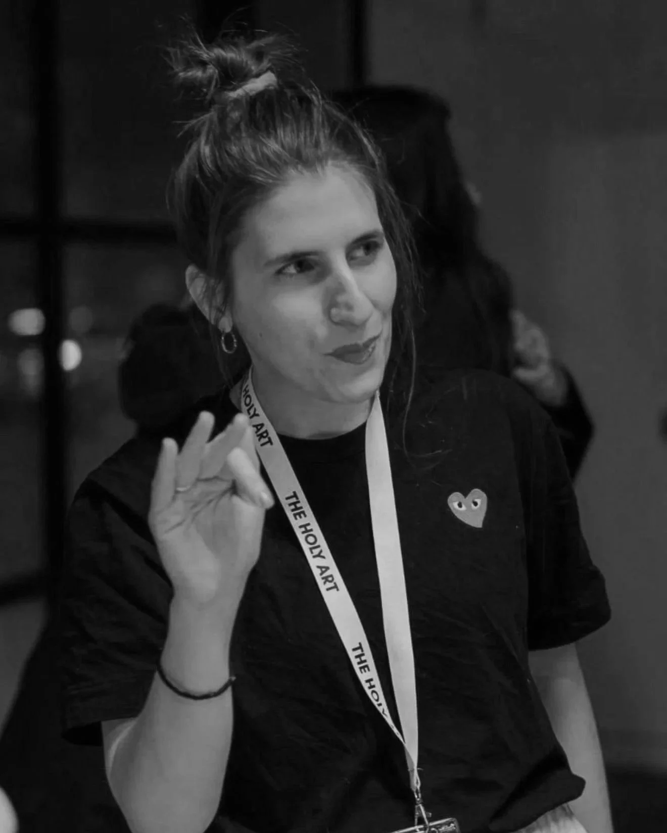 A woman with hair in a bun making an 'OK' sign with her hand, wearing a lanyard labeled 'The Holy Art' around her neck.