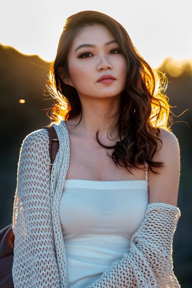 Woman with long hair wearing a white dress and a knit shawl, standing outdoors with sunlight highlighting her hair.