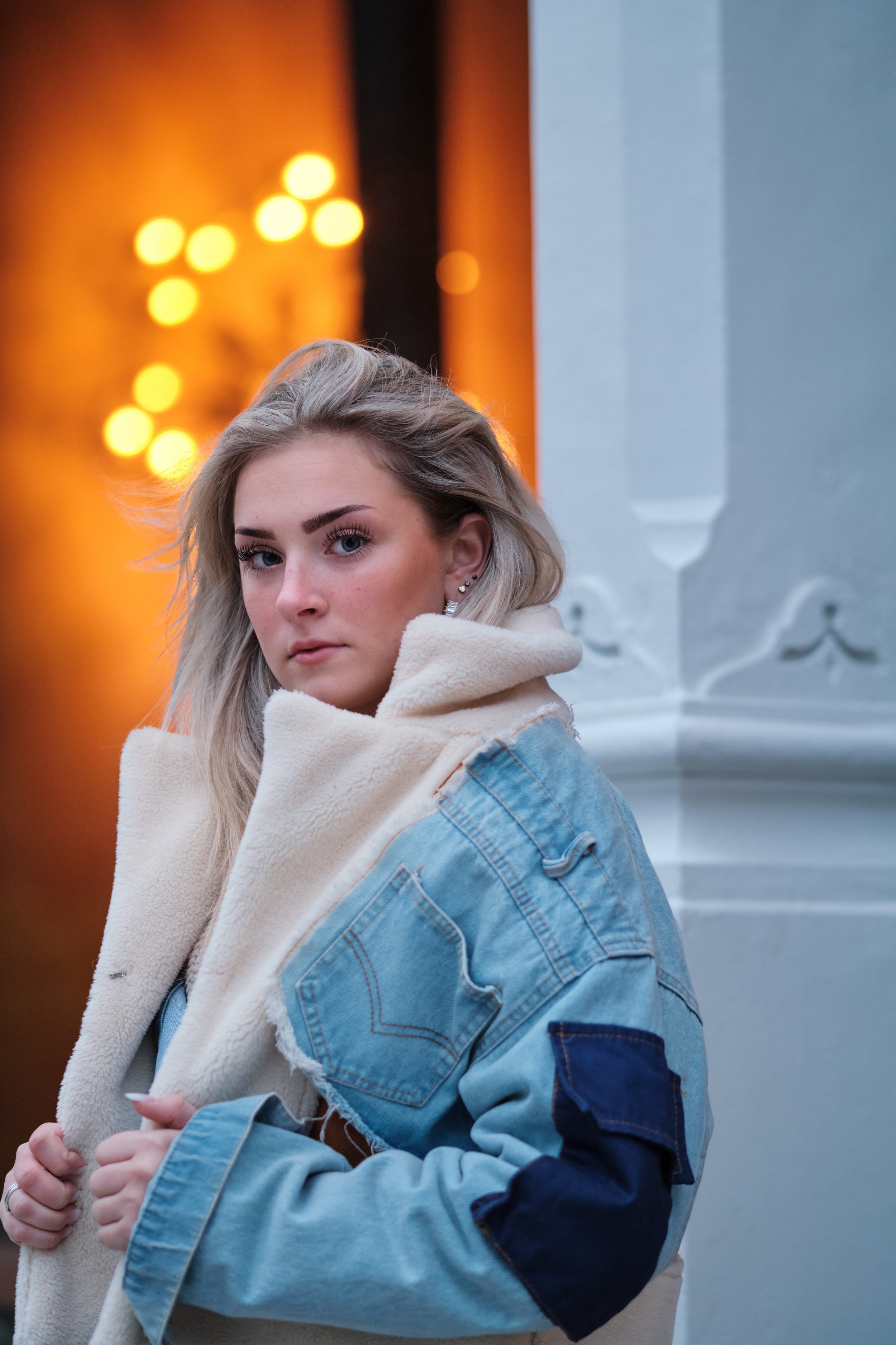 A person wearing a denim jacket with a shearling collar stands in front of a wall with orange lights.
