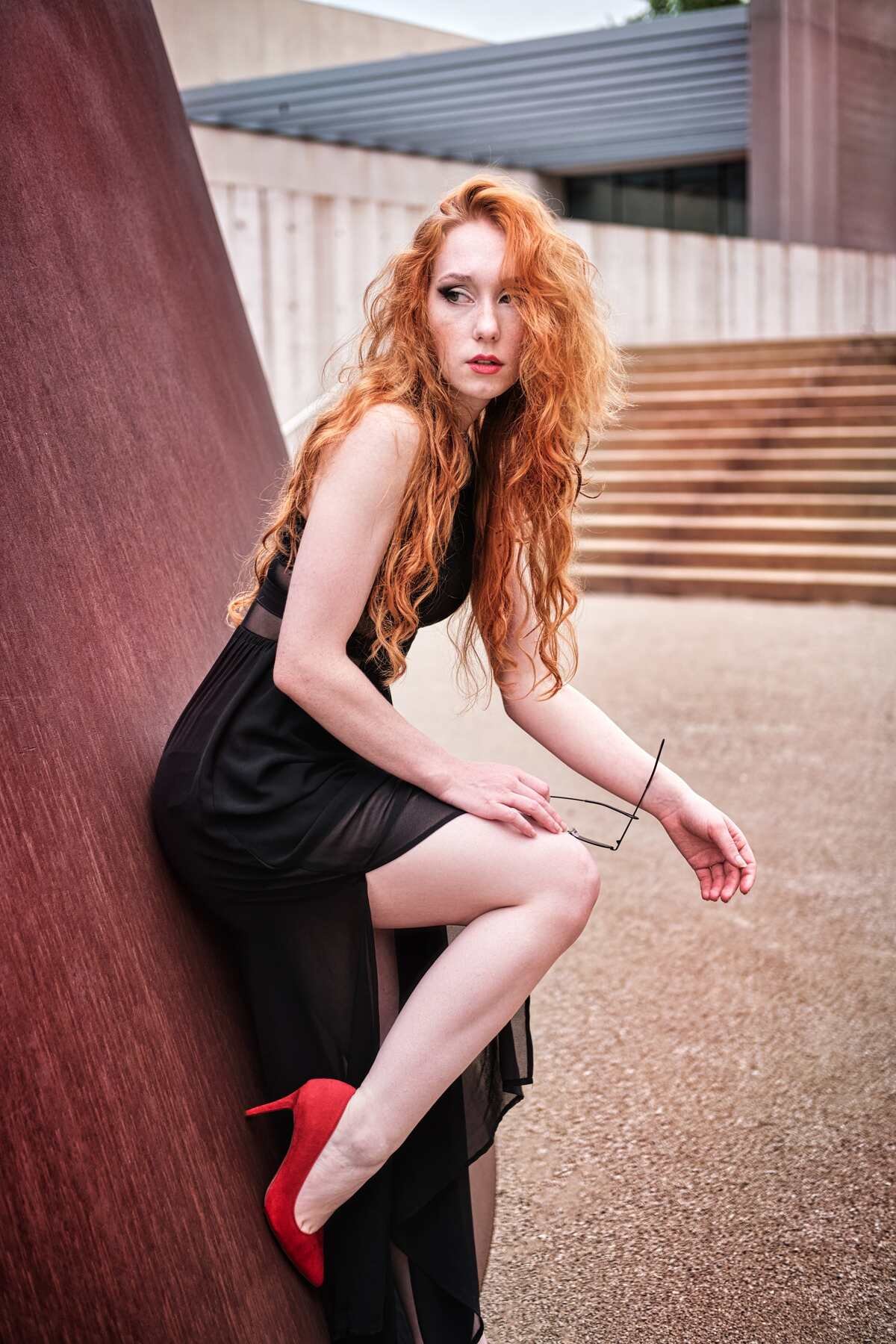 Woman with long red hair wearing a black dress and red high heels sitting on a slope, holding eyeglasses, with steps and modern architecture in the background. Photography by Henry David Photography Professional Headshots and Portraits St. Louis MO