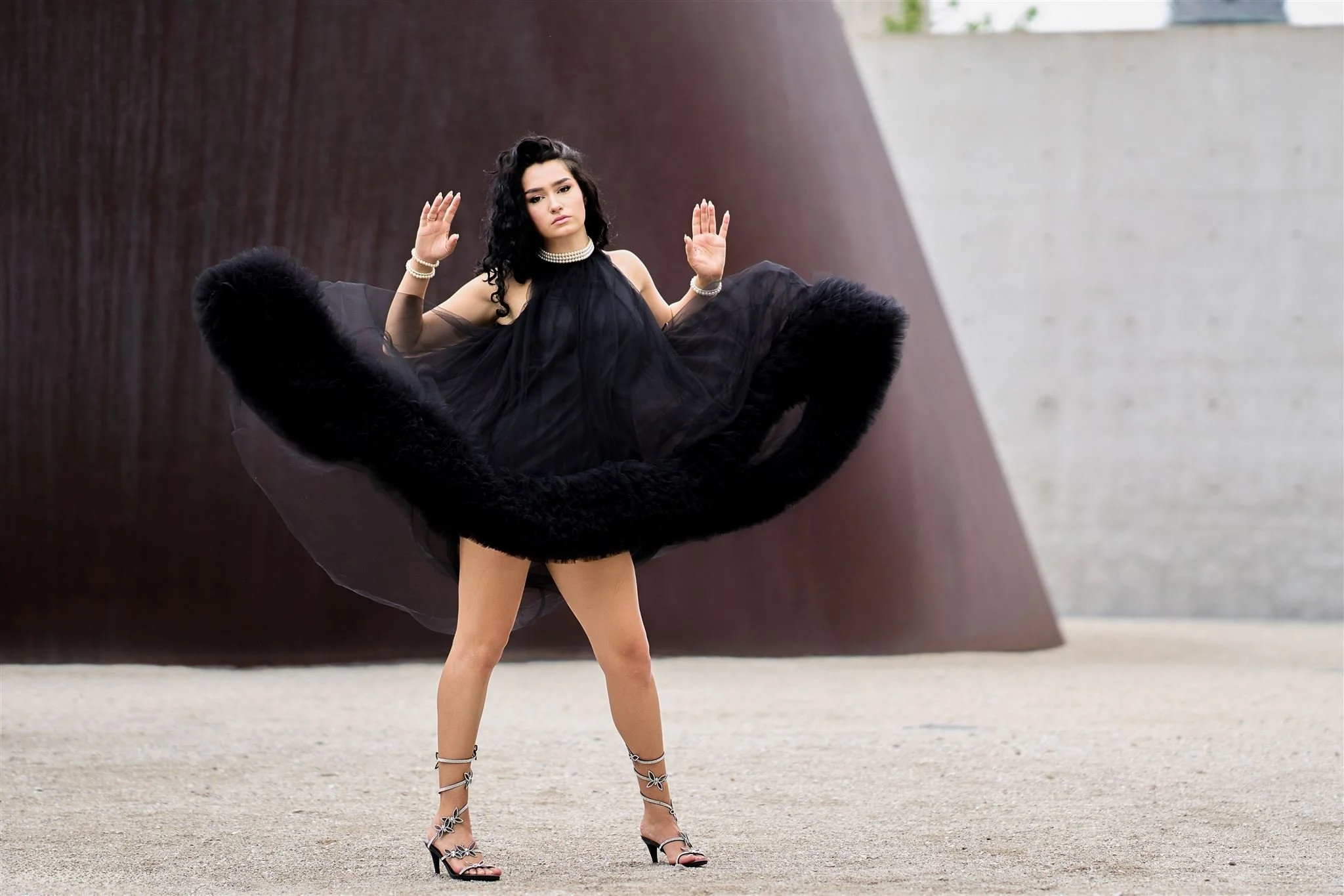 A woman with dark, curly hair wearing a black dress, high heels, and jewelry stands with arms raised in front of a modern, abstract concrete and rust-colored wall.