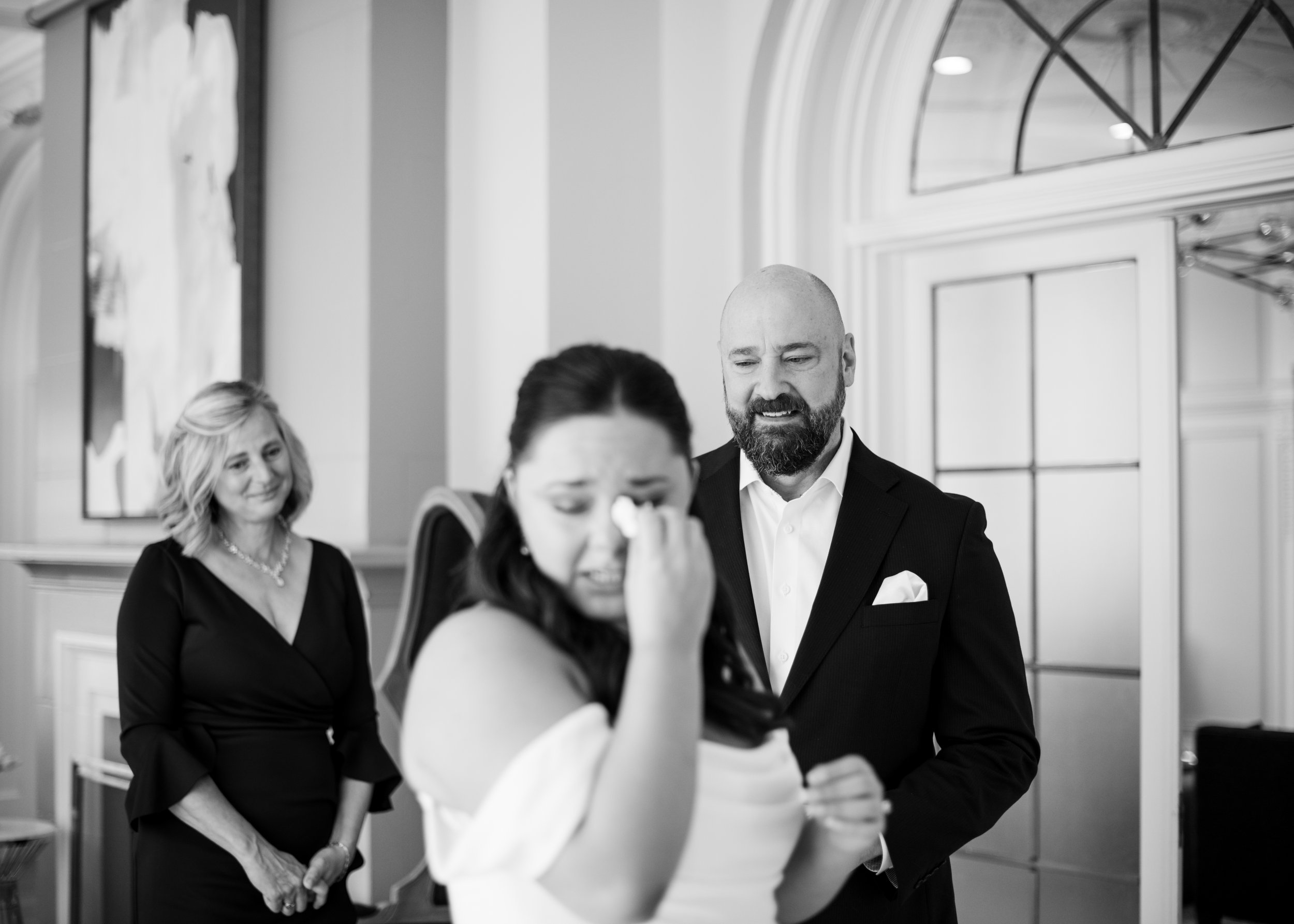 A woman in a wedding dress is crying and wiping her eye, with a man in a suit appearing emotional behind her. Another woman in a black dress stands smiling in the background.