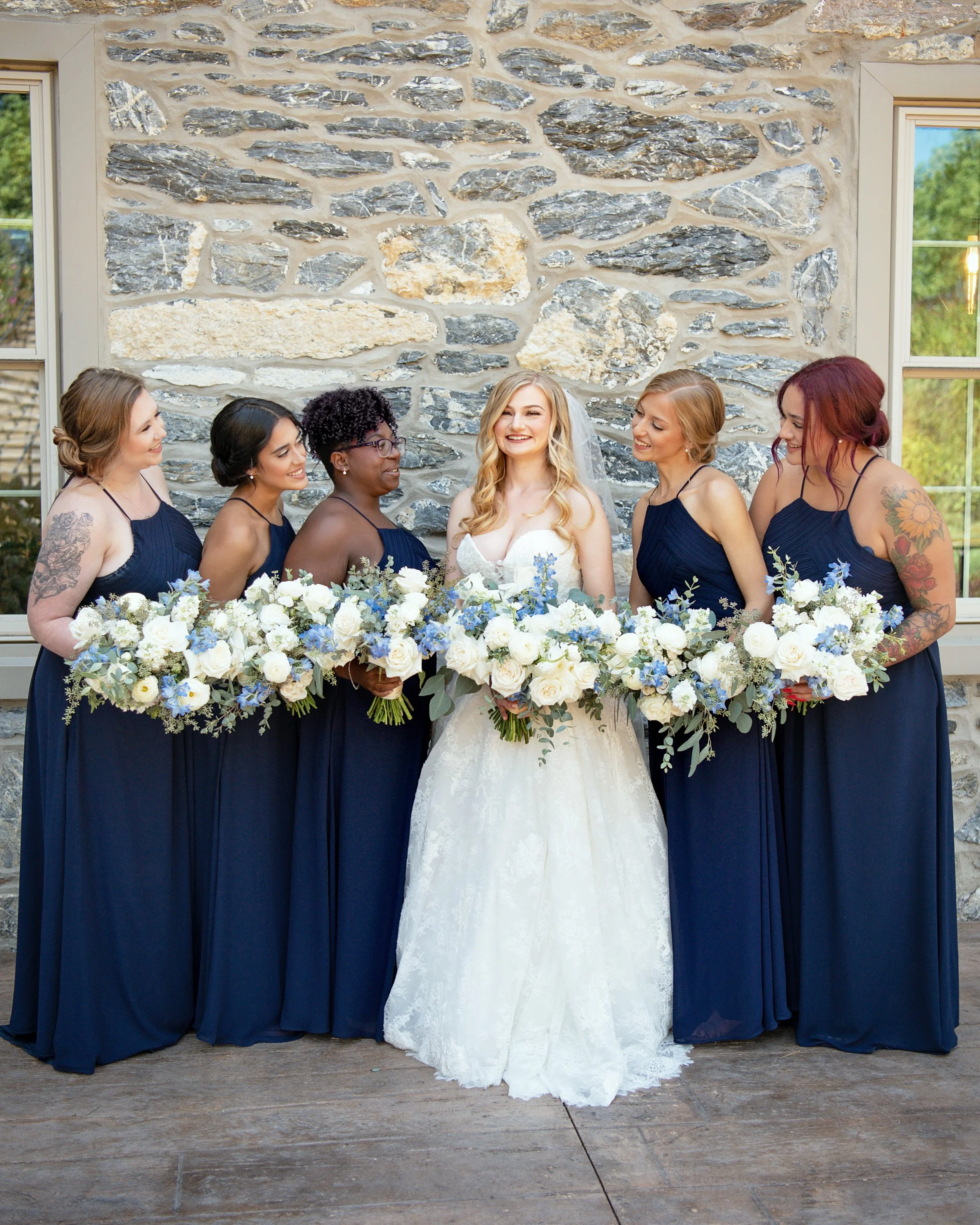 A bride in a white wedding dress standing with five bridesmaids in navy blue dresses, all holding large floral bouquets. They are posing indoors against a stone wall with windows in the background, smiling and looking at each other.