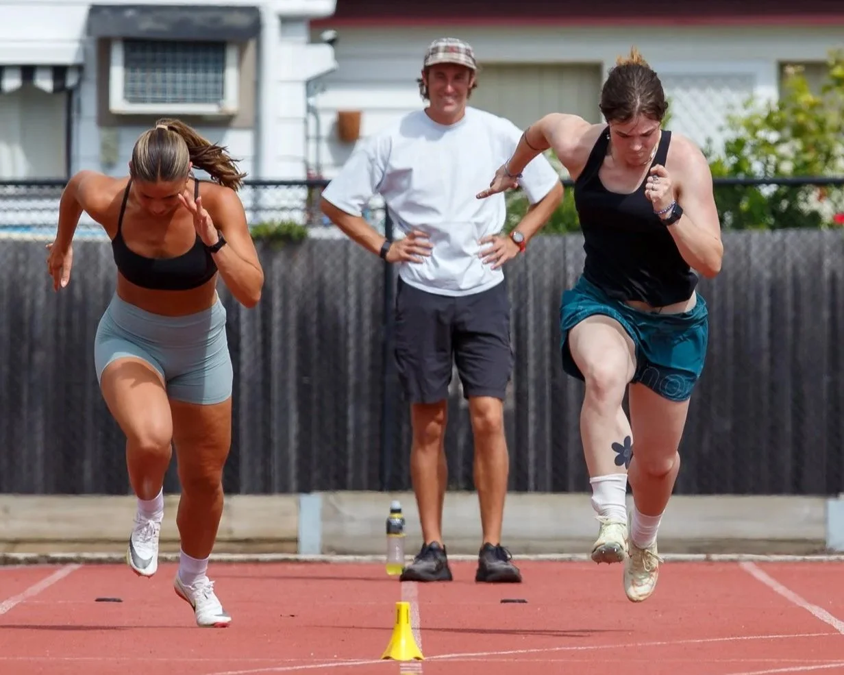 Two women sprinting on a track, with a man standing behind them watching. The woman on the left wears a black sports bra and light shorts; the woman on the right wears a black tank top and teal shorts. They are competing in a race, and there are small markers on the track.