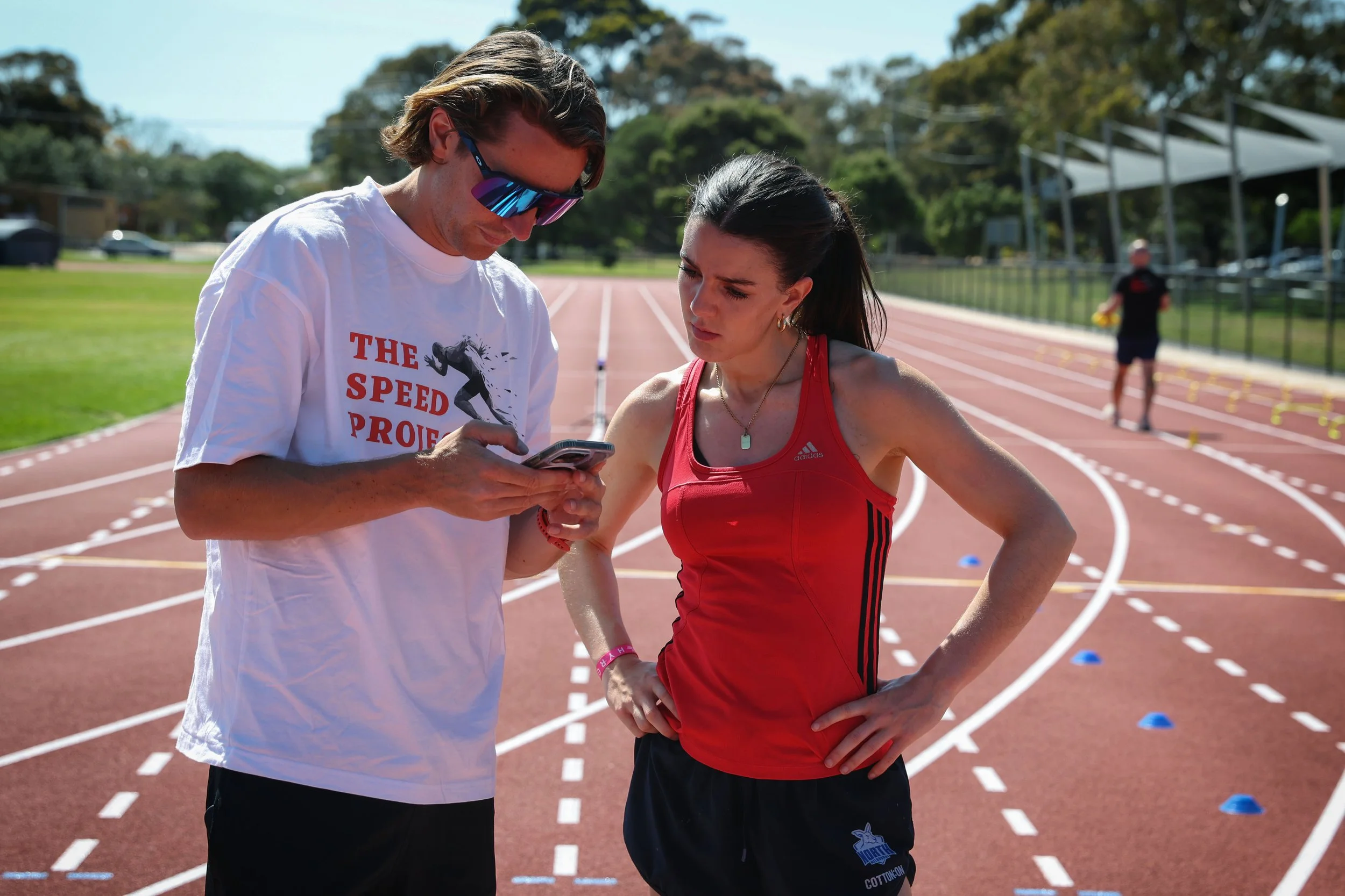 A male coach and a female athlete are standing on a running track, looking at a phone together during daytime. The coach is wearing a white T-shirt that says 'The Speed Project' and sunglasses, while the athlete is wearing a red athletic tank top and black shorts. There are other athletes in the background, and the scene is set outdoors with trees and a clear sky.