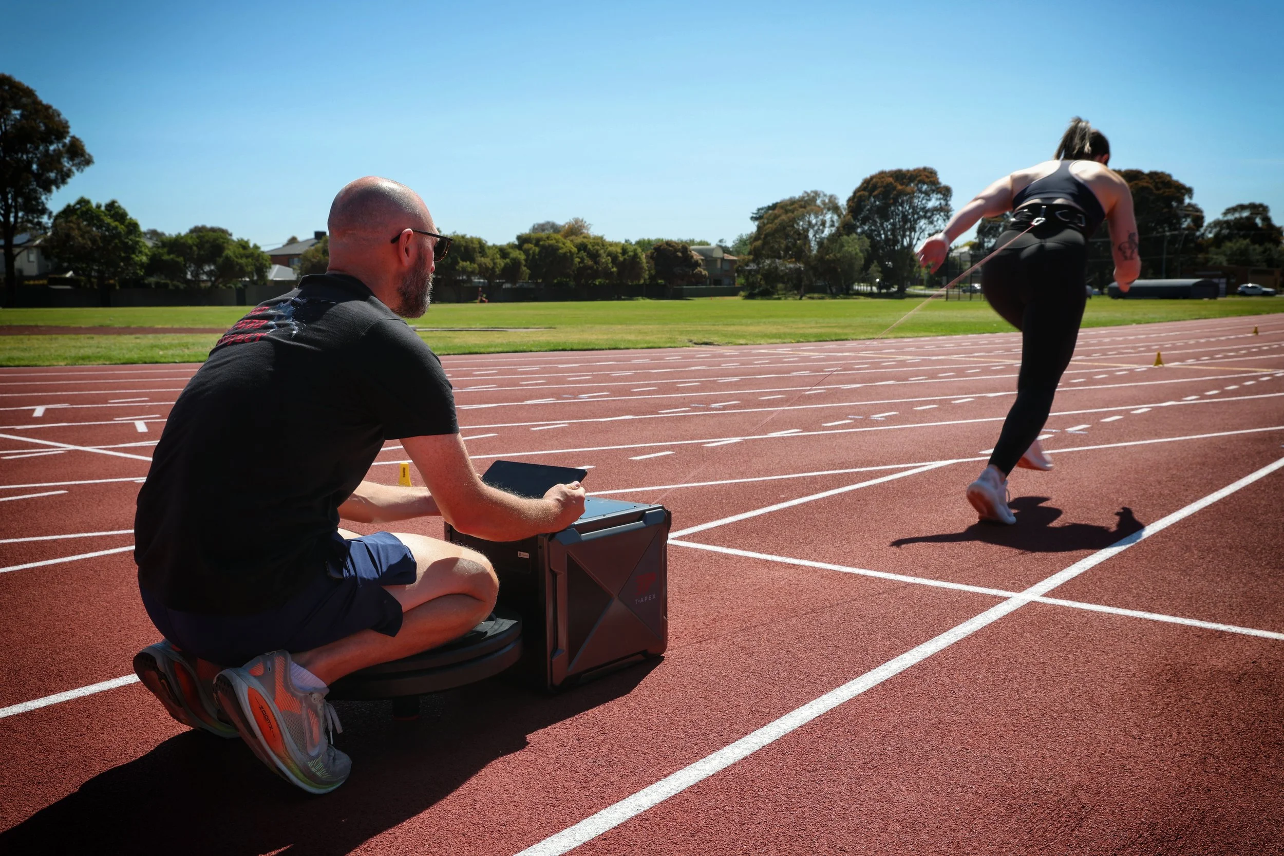 A female sprinter starting a race on a track, with a coach or technician kneeling nearby, monitoring her performance with equipment, under a clear blue sky with trees in the background.