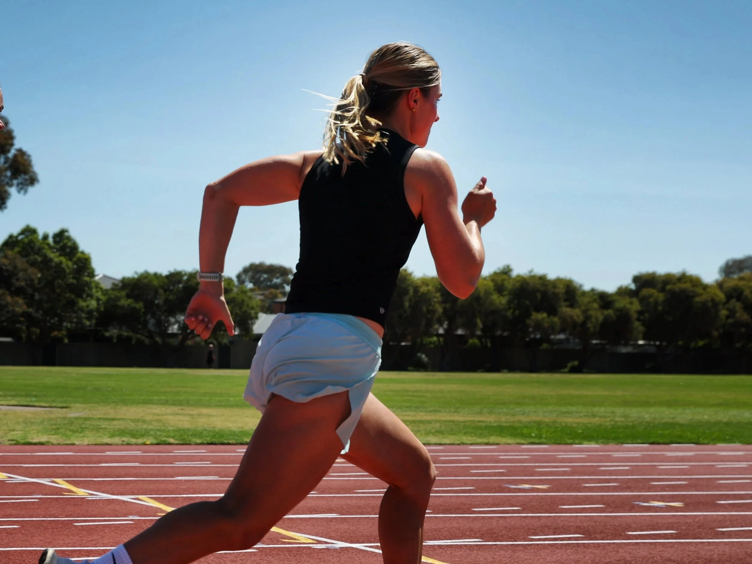A woman running on a track in an outdoor stadium on a sunny day, wearing a black sleeveless top and light-colored shorts.