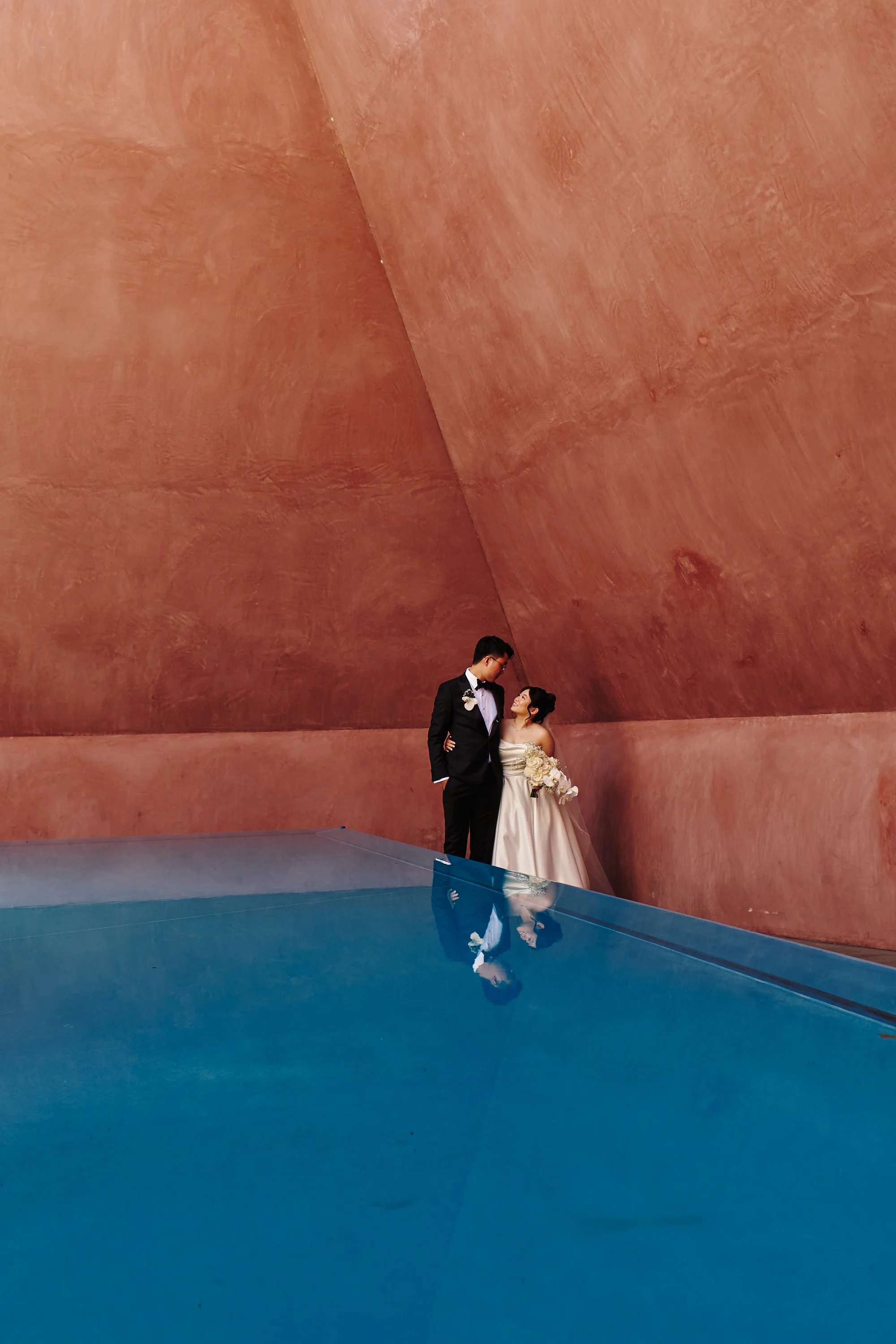 A bride and groom standing close together in a modern architectural setting with large pinkish-orange walls. The groom is wearing a black tuxedo and the bride is in a white wedding gown holding a bouquet. Their reflection can be seen on a shiny blue 