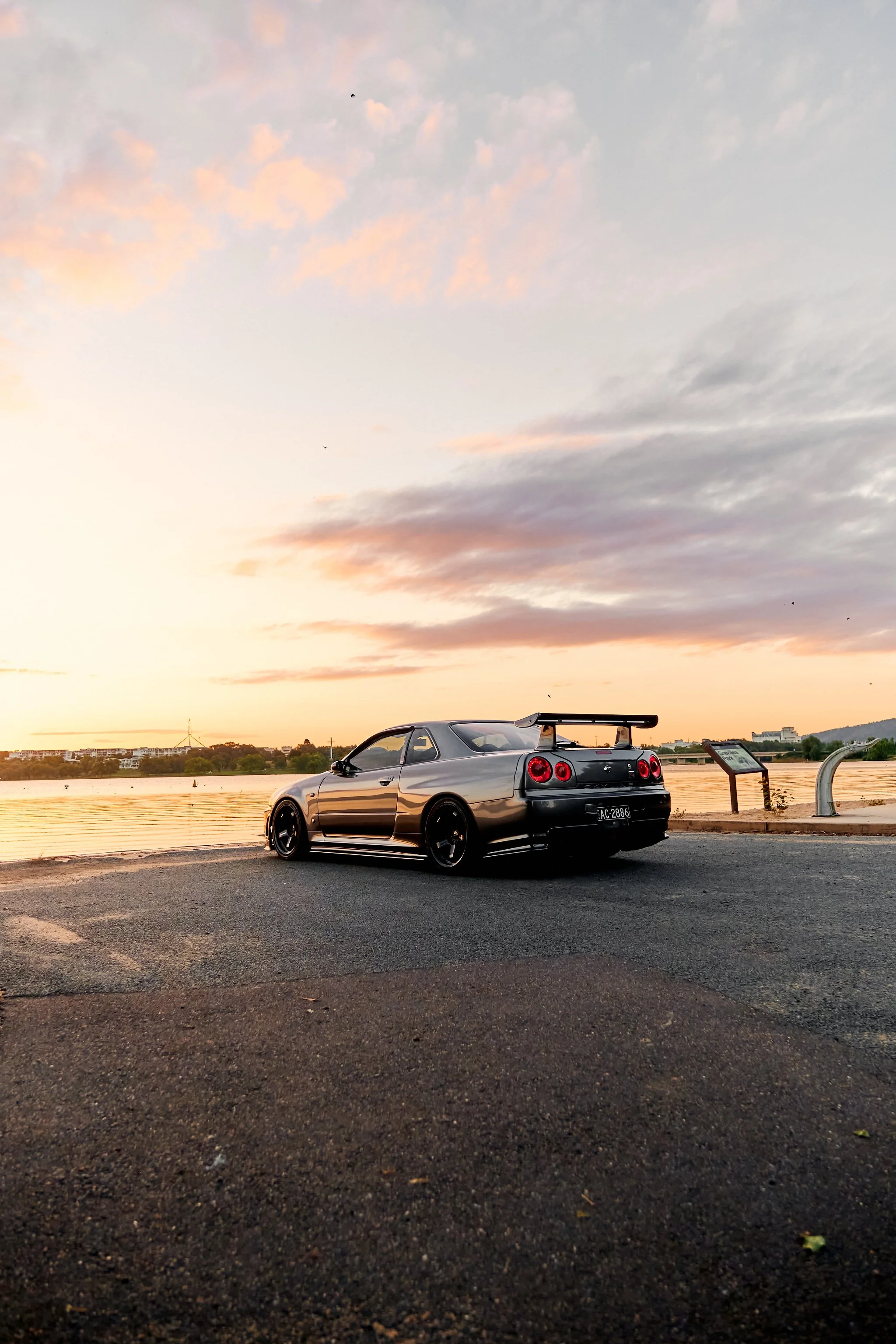 A black sports car parked near a body of water during sunset, with colorful clouds in the sky and a cityscape in the background.