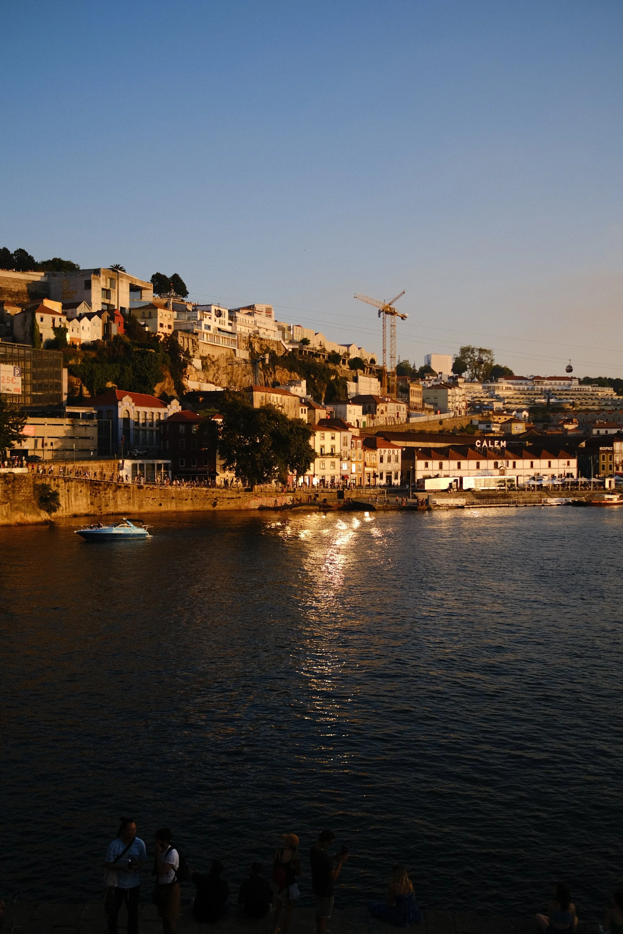 People standing by a river at sunset with a hillside cityscape, construction crane, and a cable car in the background.