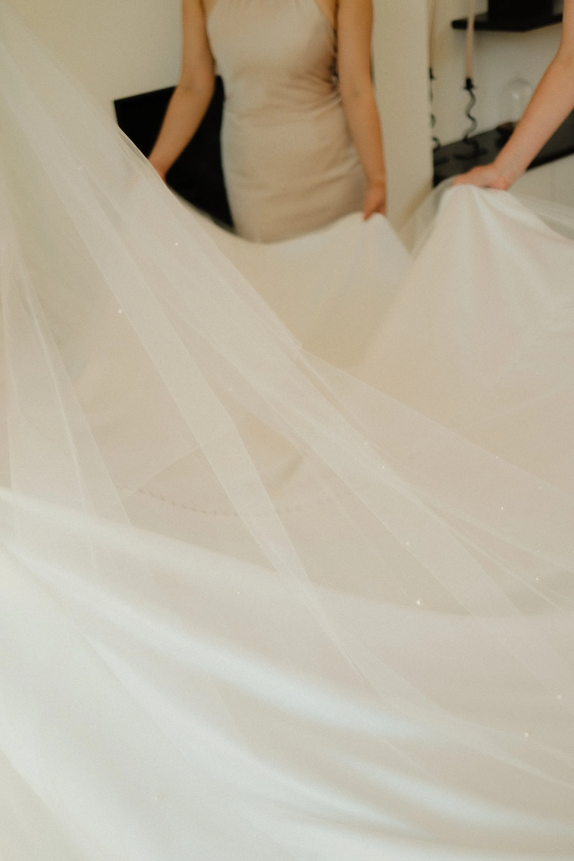 Part of an indoor scene showing a woman in a light-colored dress standing near a white fabric, with other people and furniture in the background.