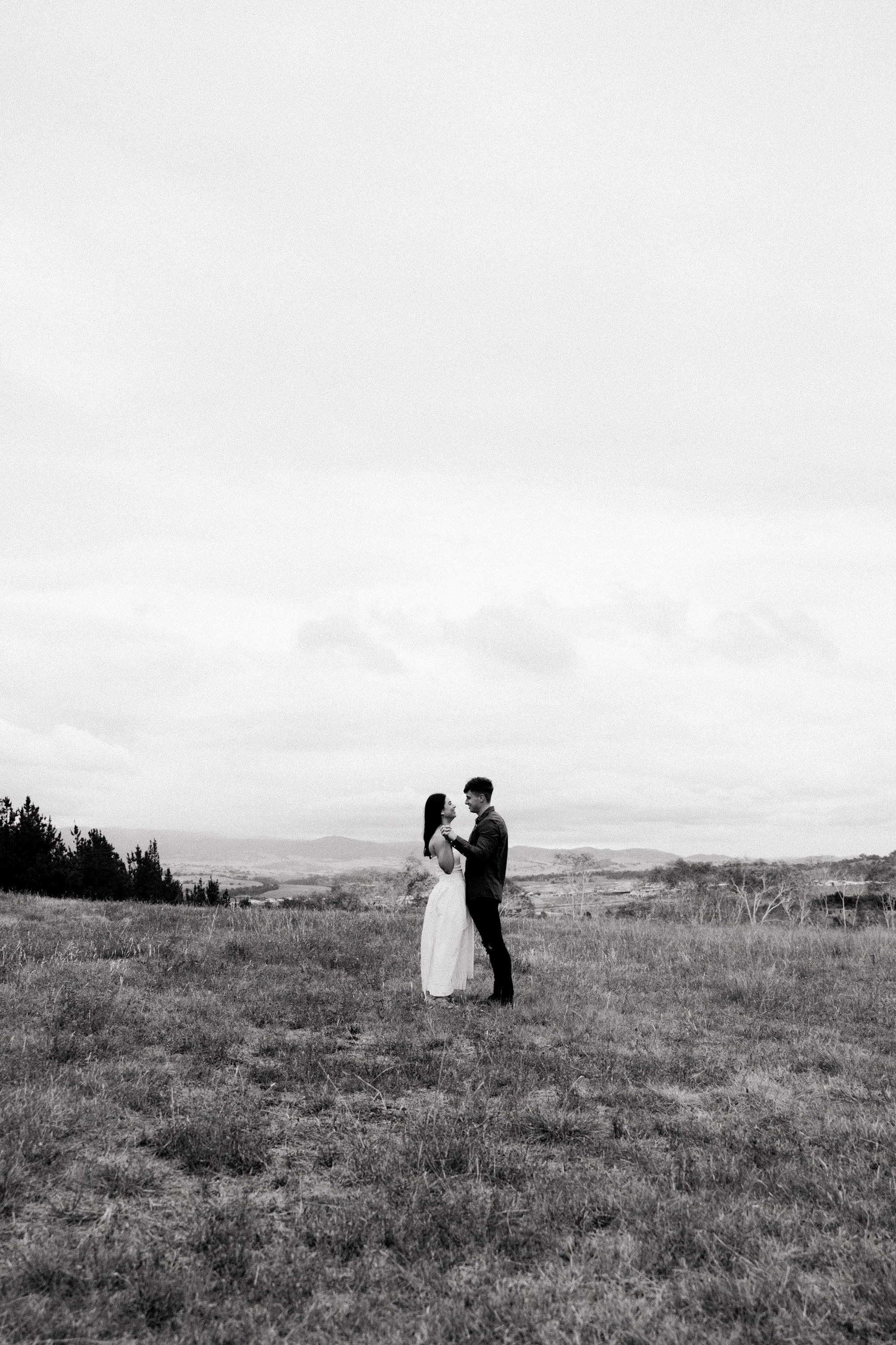 A black-and-white photo of a couple standing in a grassy field, holding hands and looking into each other's eyes. The woman is wearing a long dress, and the man is in dark clothing. The landscape in the background includes rolling hills and sparse tr