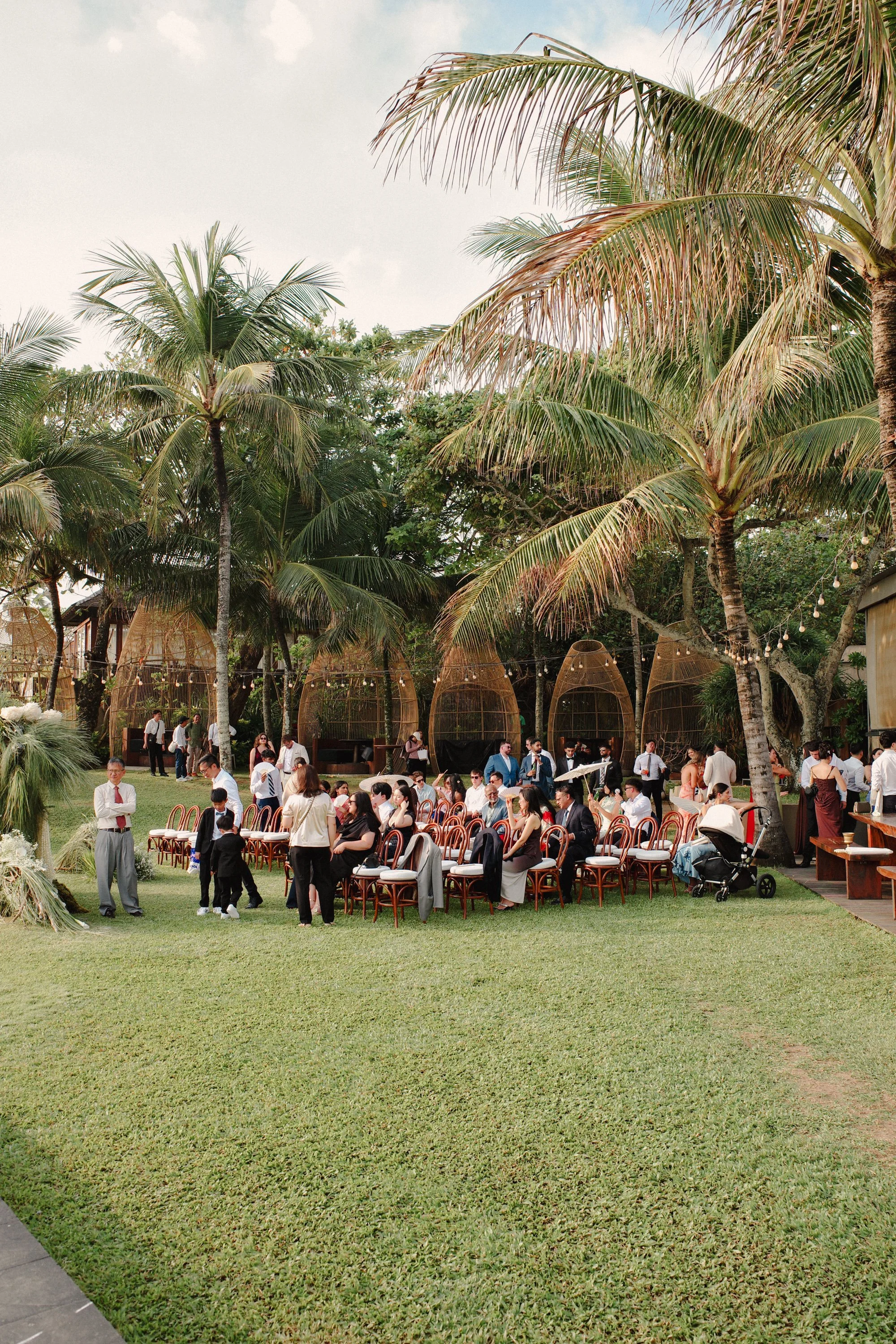 An outdoor gathering with numerous people seated and standing on a well-maintained lawn, surrounded by tall palm trees and tropical foliage, with decorative wicker structures and string lights in the background.