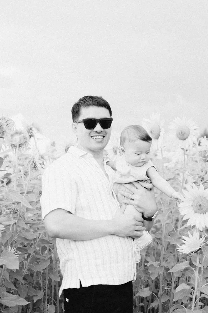 Man with sunglasses holding a baby girl in a sunflower field