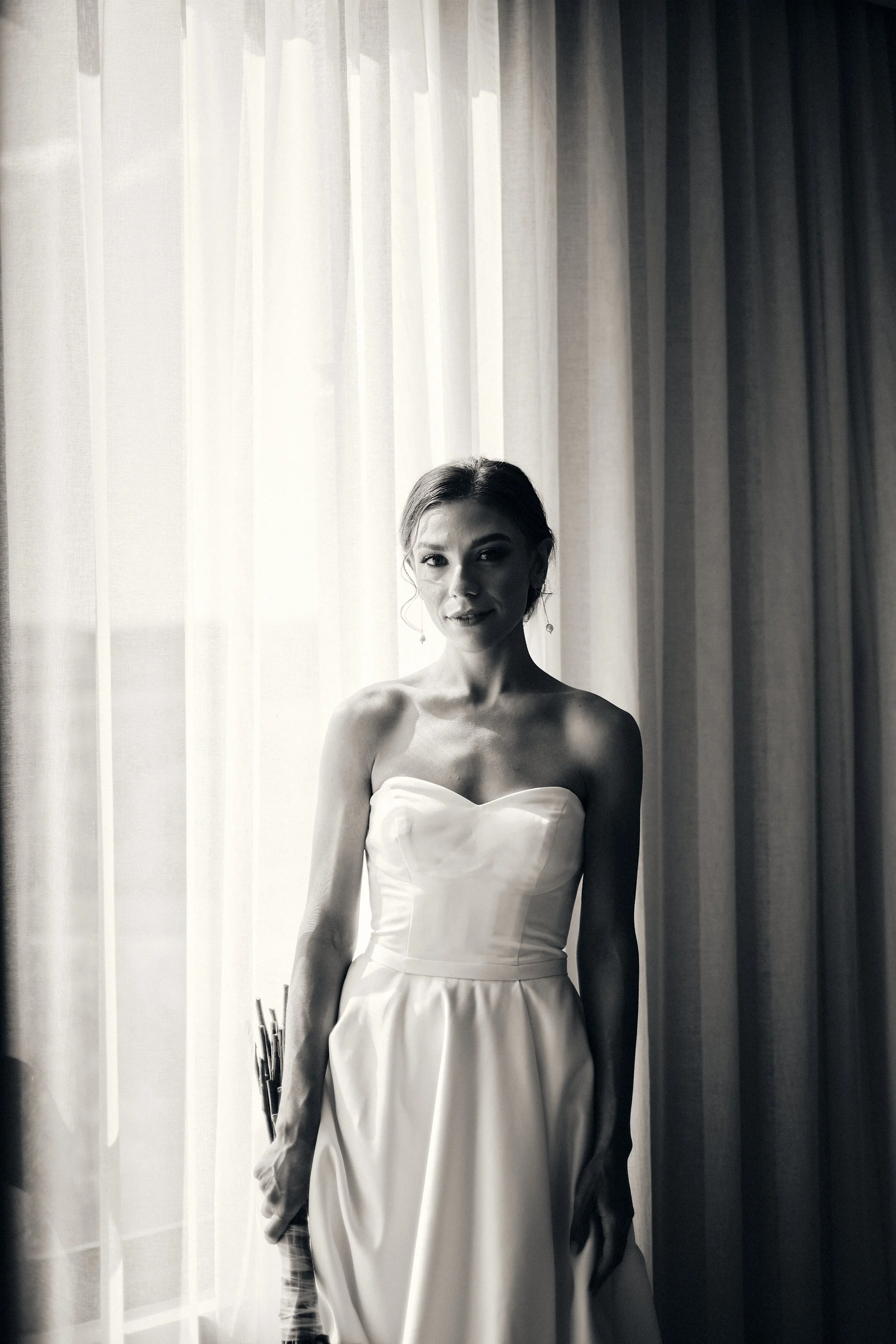 A woman in a strapless white dress standing by sheer curtains with sunlight filtering through, in a black and white photograph.