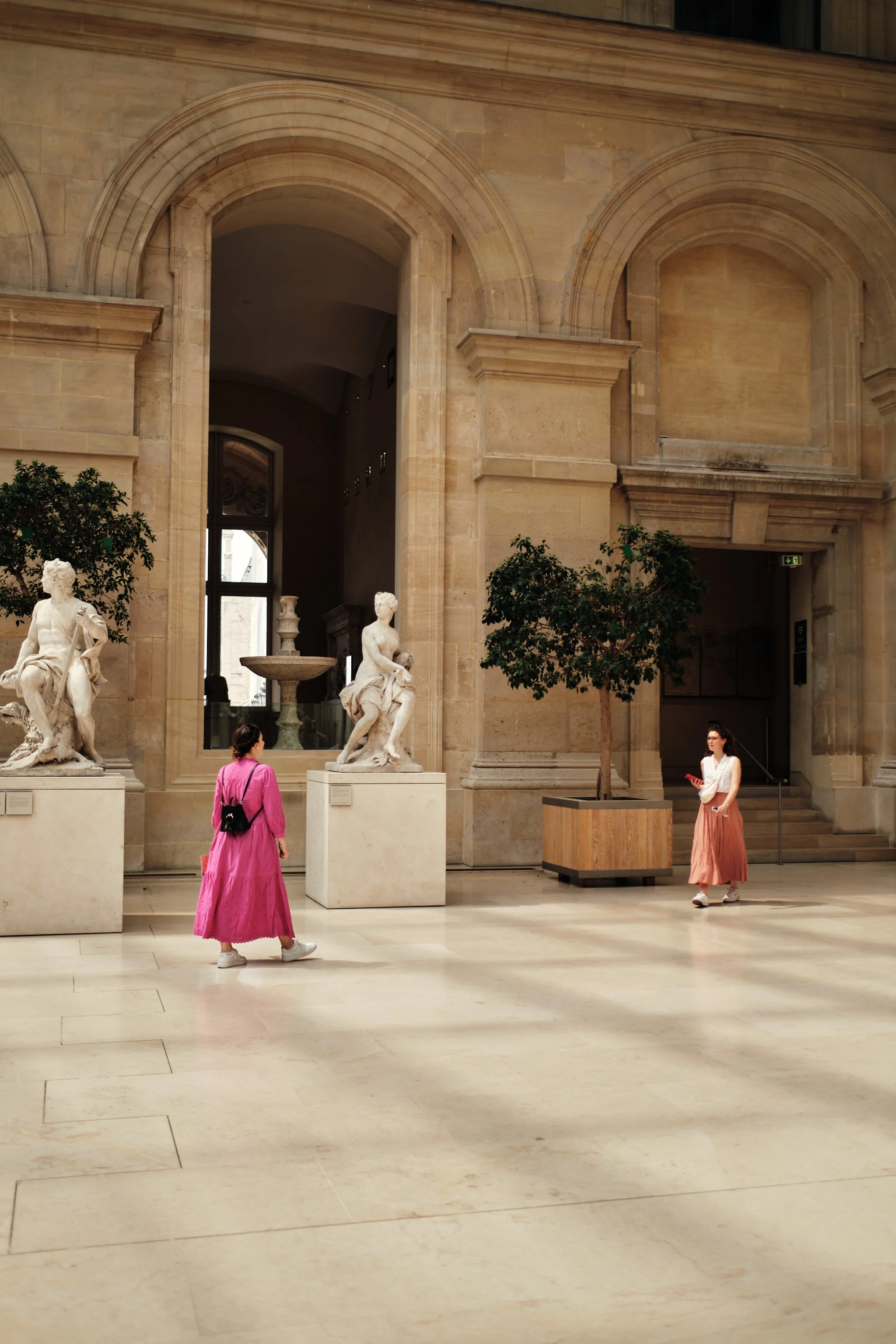 Museum interior with classical statues on pedestals, two women walking and standing, modern planters with small trees, and large arched windows in the background.