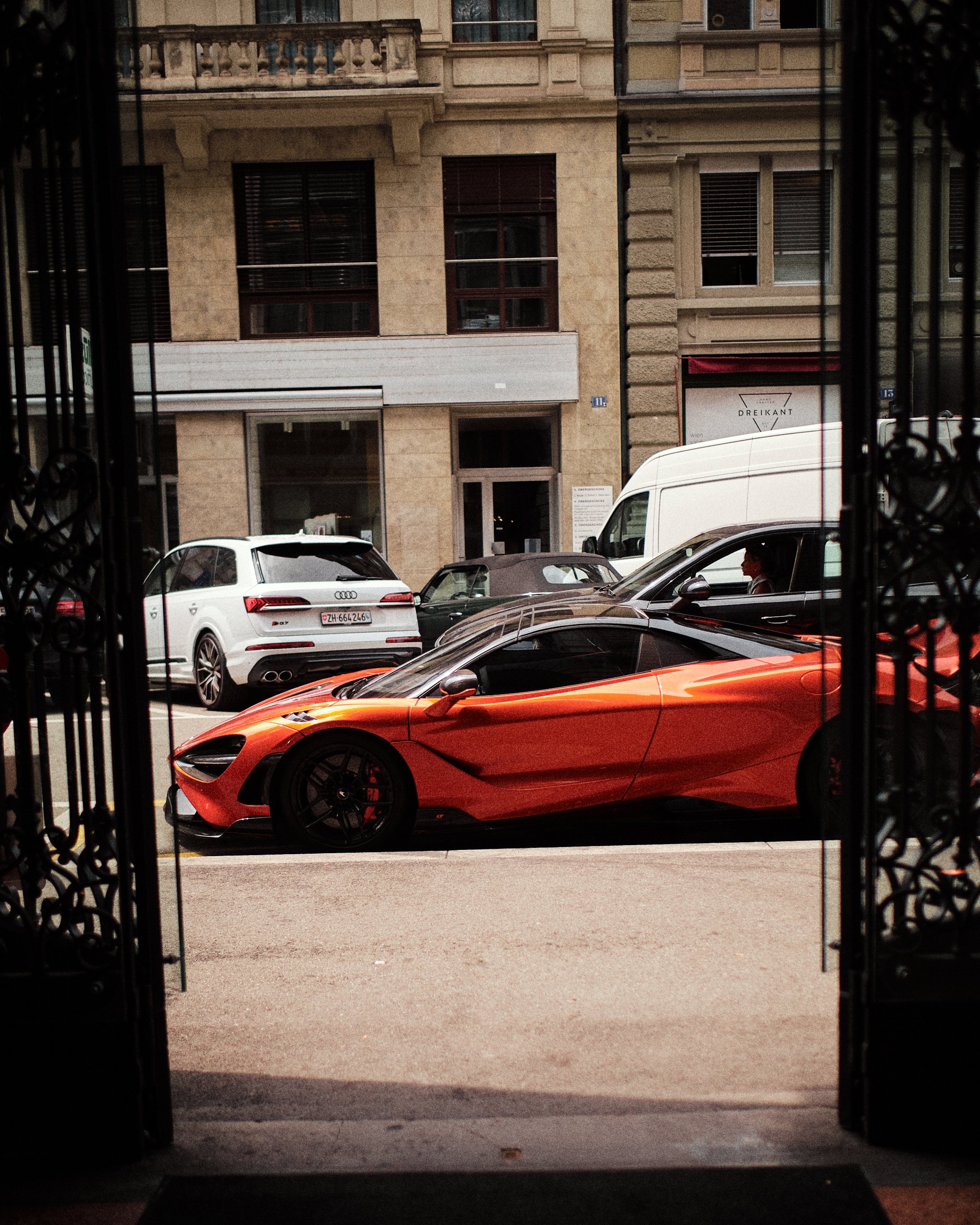 View of parked cars on a city street seen through an open gate. A luxury orange sports car is in the foreground, with a white Audi and a white van further back. The street is lined with old buildings featuring large windows and balconies.
