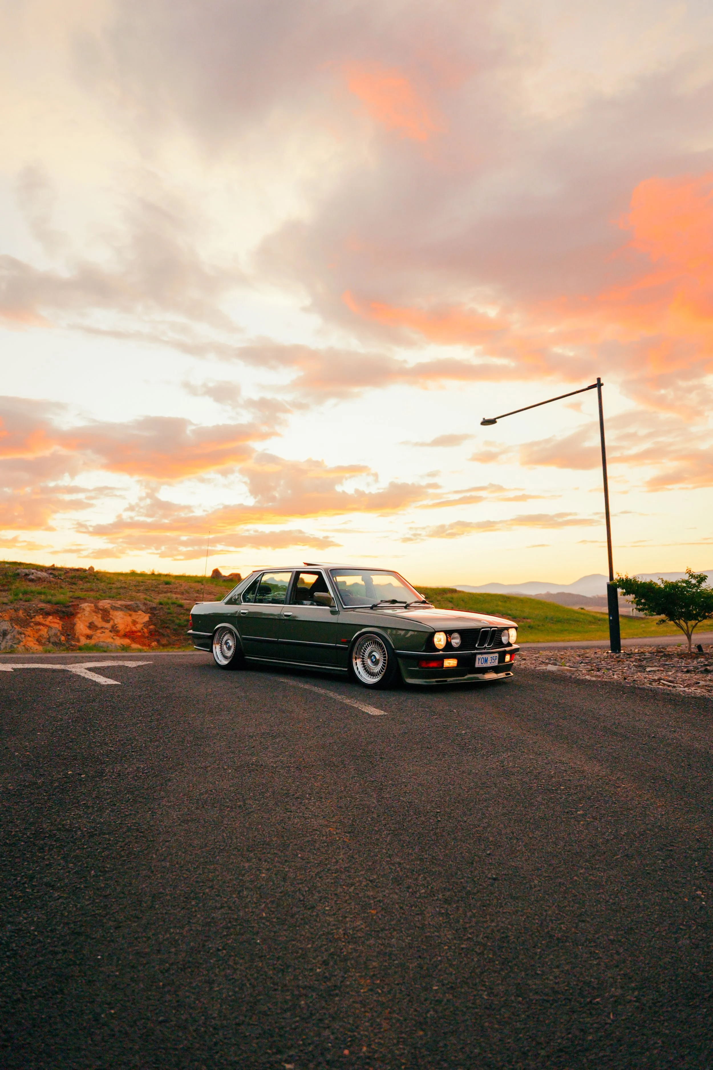 A black vintage BMW car is parked on the side of a highway at sunset. The sky is filled with orange, pink, and gray clouds, and there is a streetlamp to the right of the car. The landscape includes grassy hills and mountains in the distance.