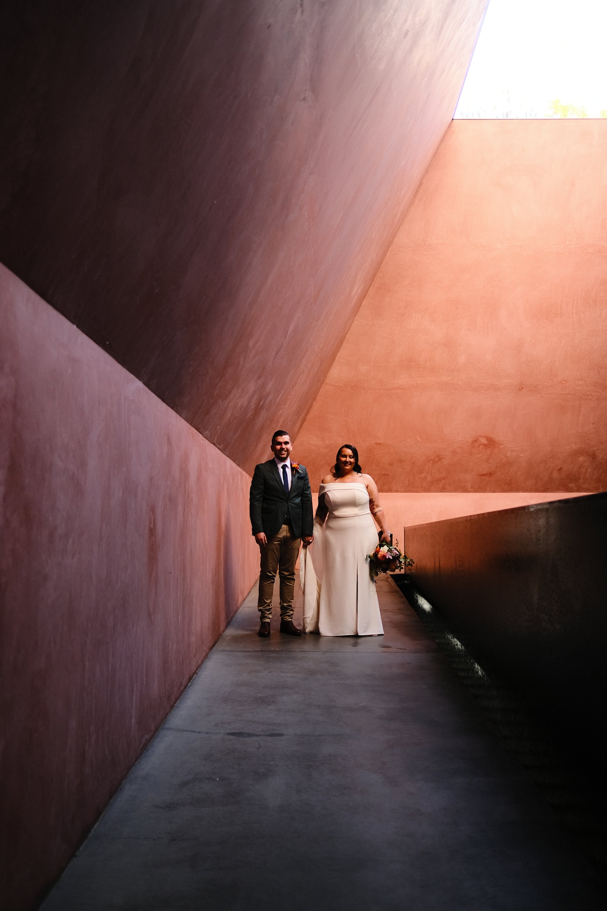 A bride and groom standing together in a modern architectural setting with pink and brown walls and an overhanging roof.
