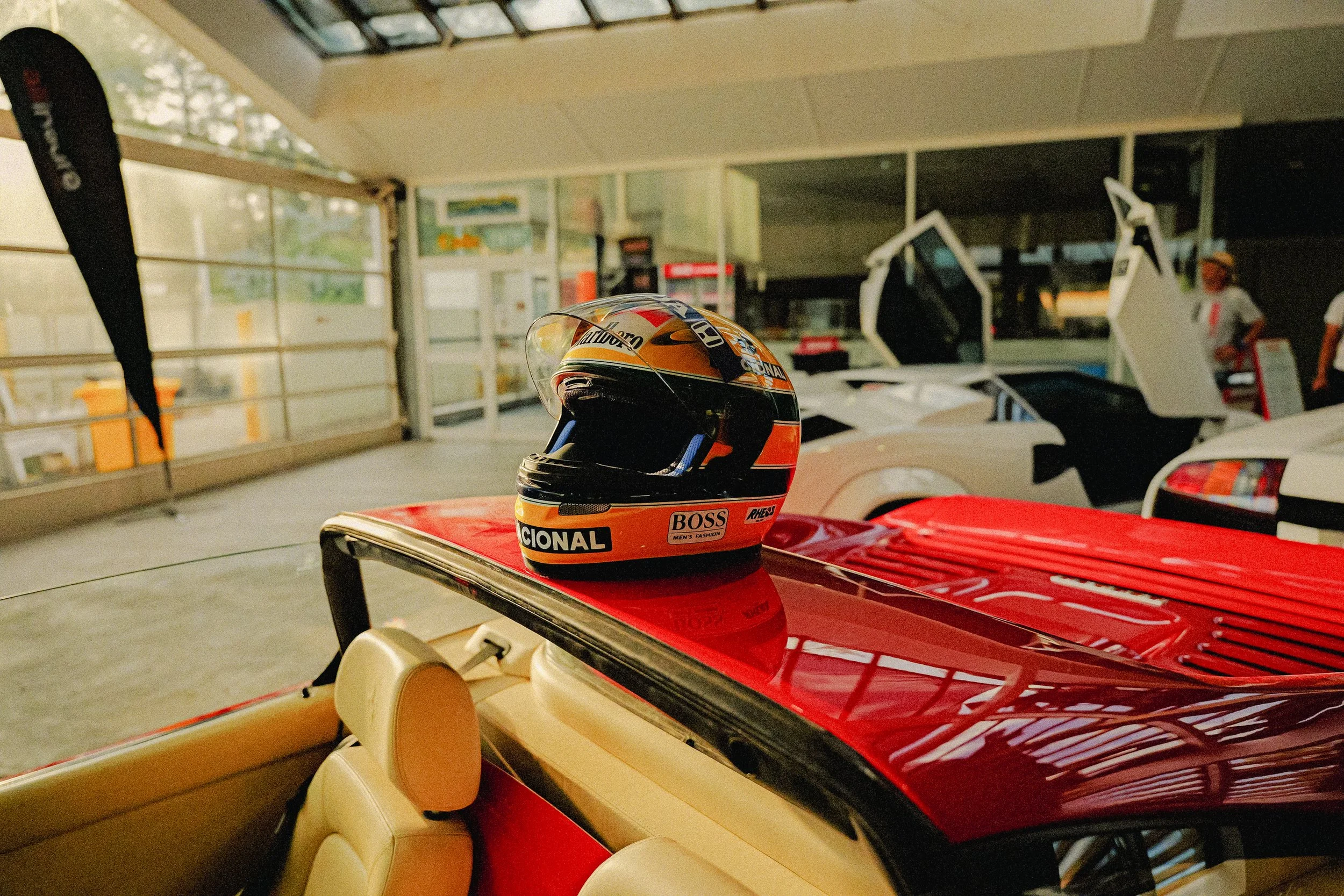 A racing helmet placed on a red race car hood inside a garage, with other cars and a person in the background.