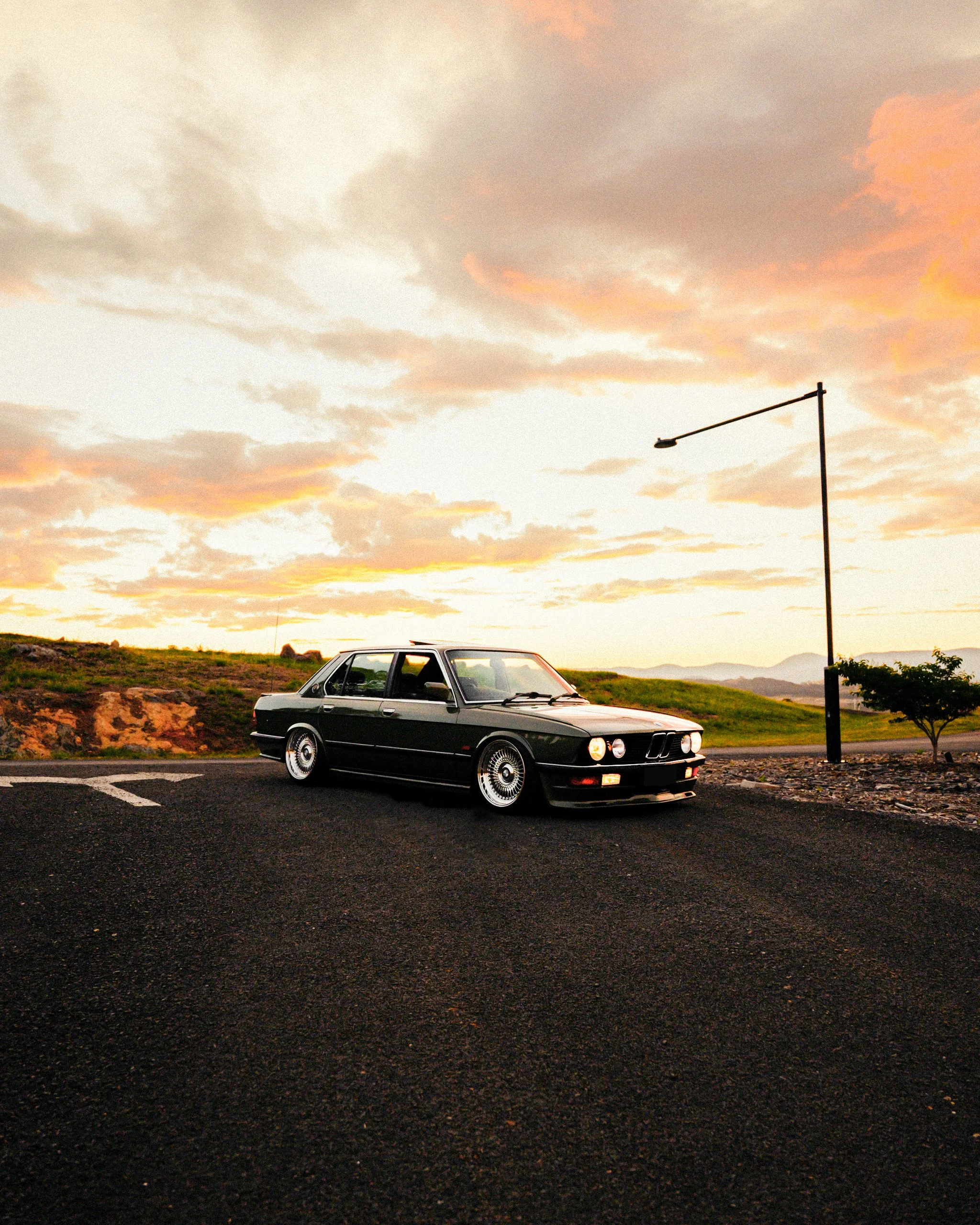 A black vintage car parked on a dark asphalt road during sunset with a cloudy sky and a streetlamp in the background.