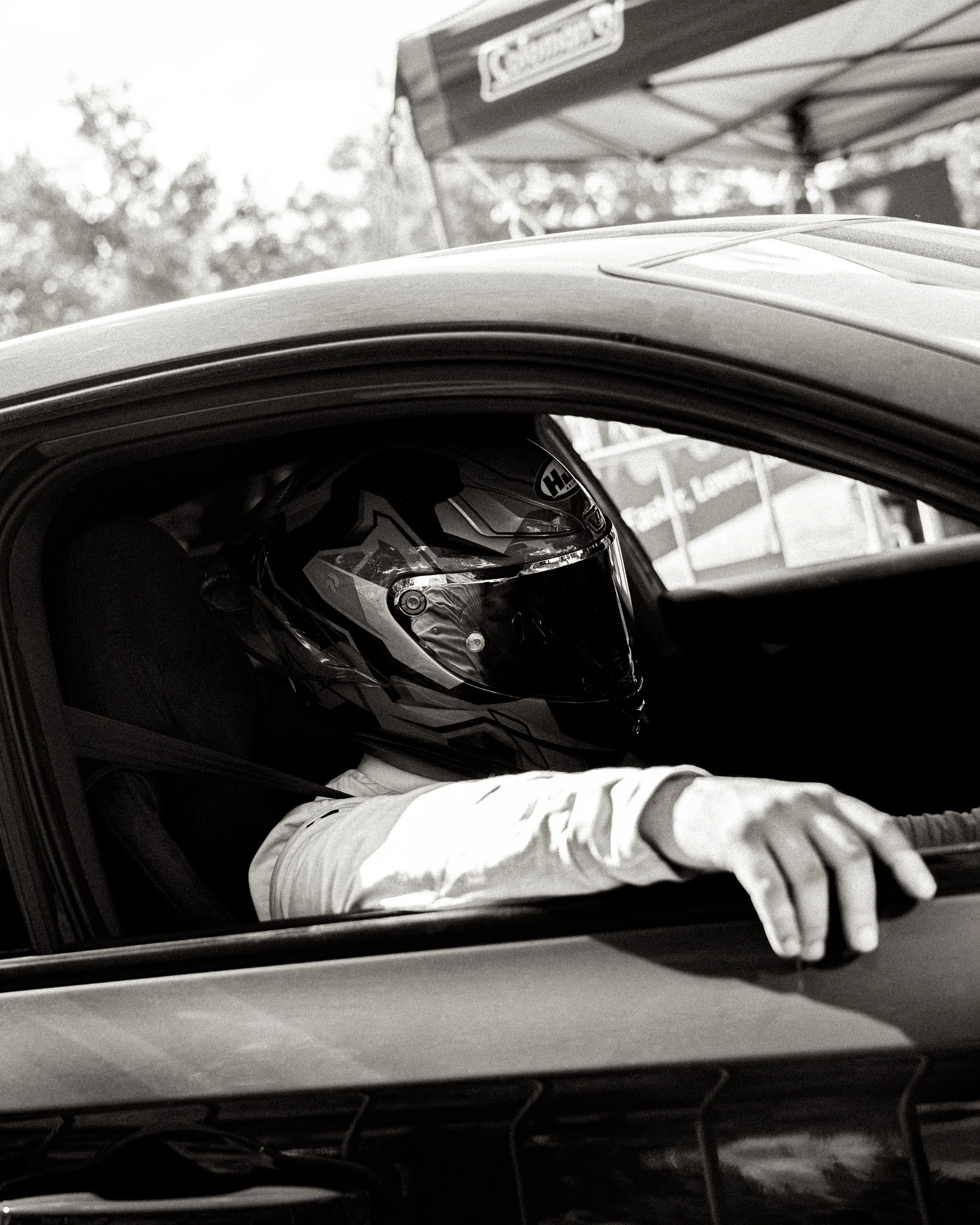A person lying on a stretcher with a motorcycle helmet covering their face. The image is black and white, taken from outside a vehicle, with an out-of-focus background showing trees and a sign.