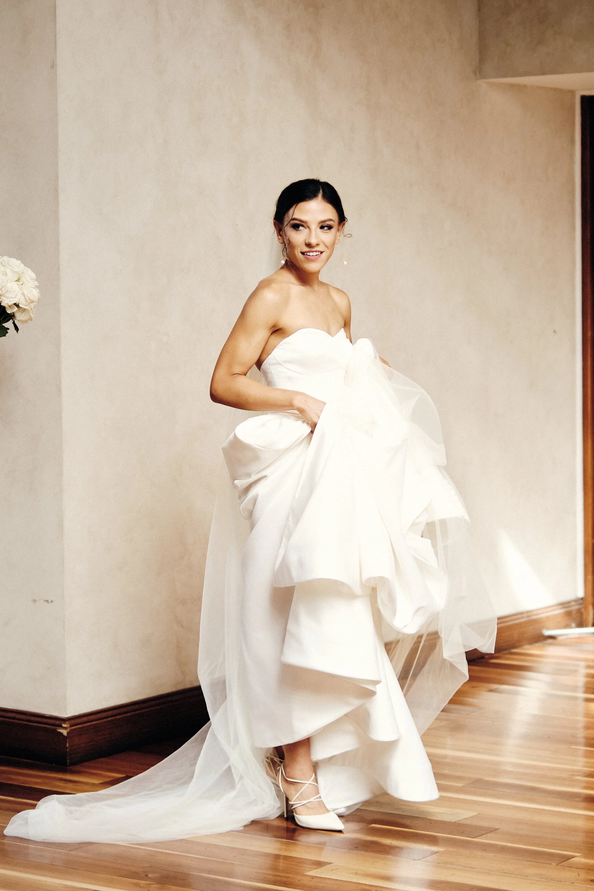 A woman in a strapless white wedding dress with a long train, standing on a wooden floor indoors, holding the fabric of her dress, with dark hair and earrings, smiling.