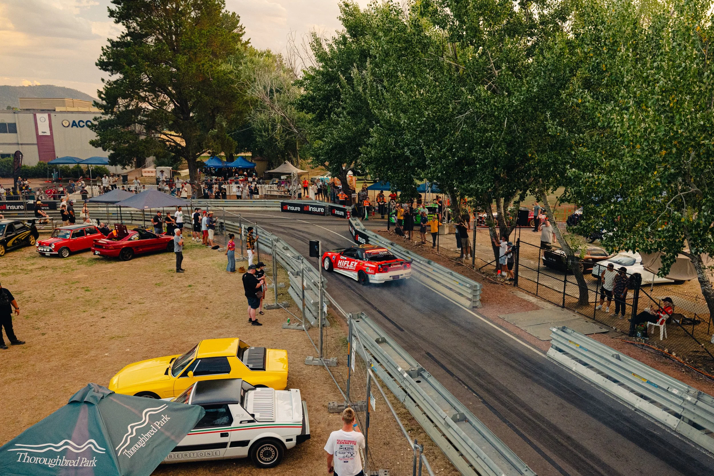 A racetrack with race cars, spectators, and trees. Classic cars and tents are visible at the foreground. The scene shows a motorsport event at Thoroughbred Park.