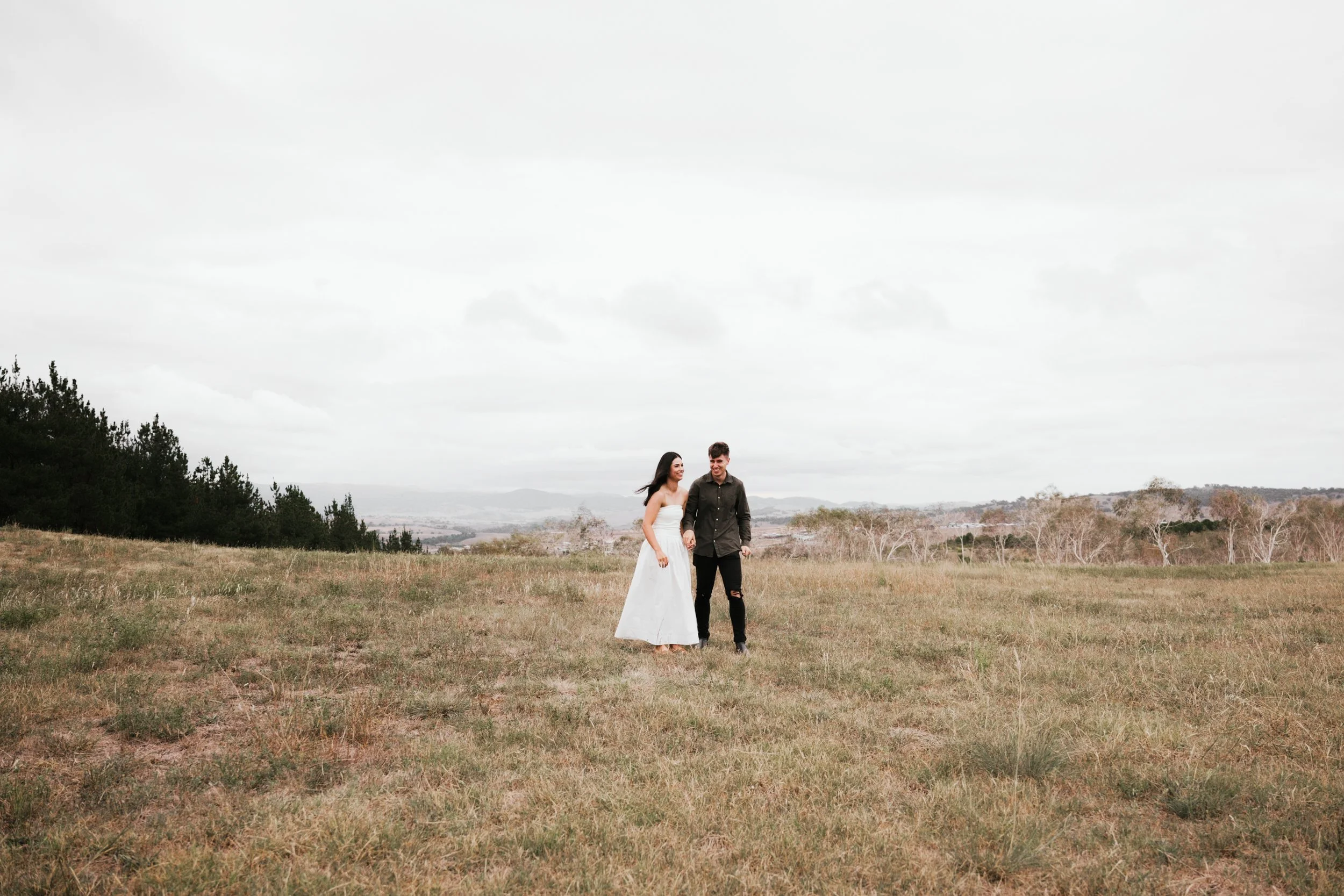 A couple walking hand in hand in an open grassy field on an overcast day, with trees and distant hills in the background.