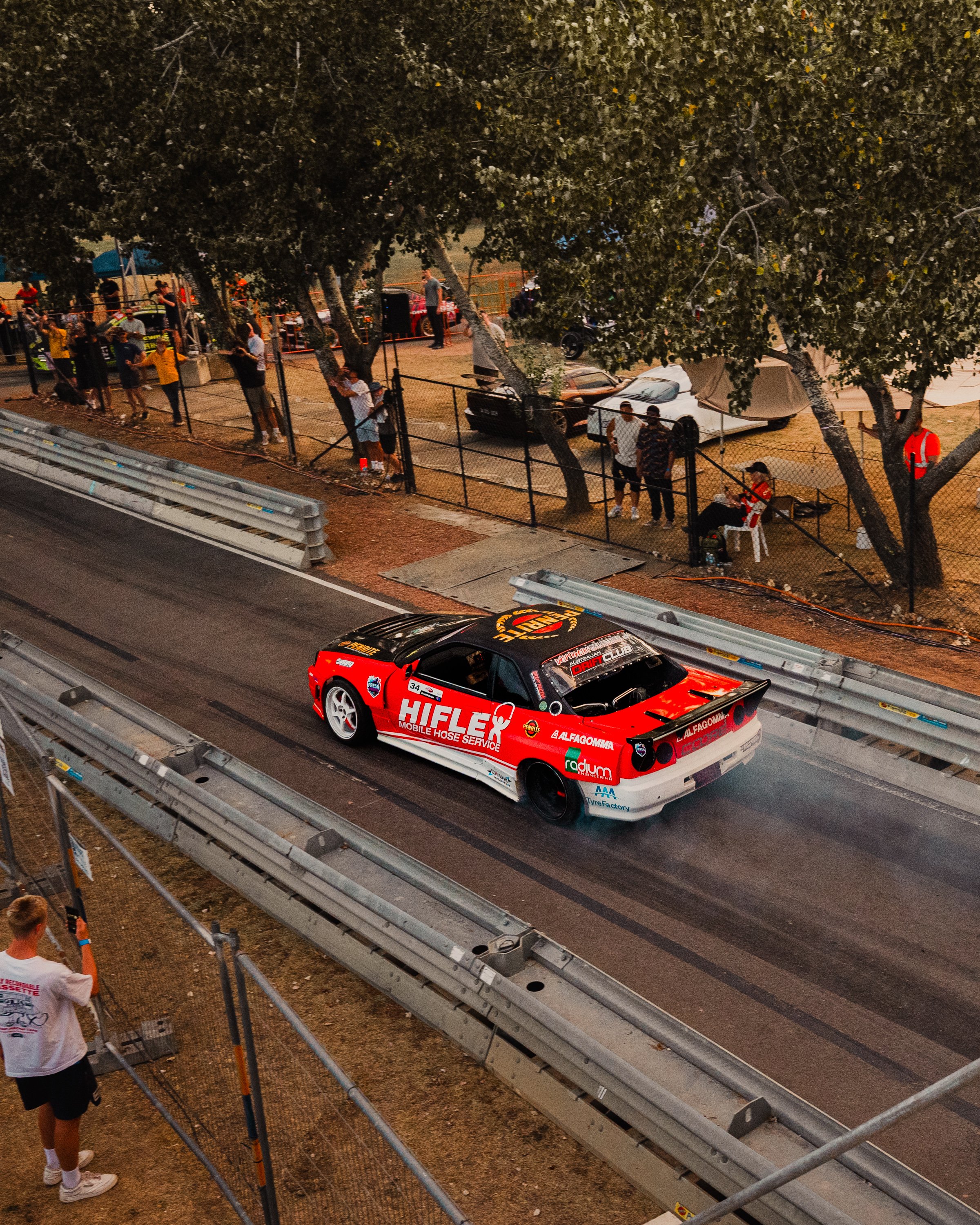 A red race car on a racetrack with the number 34, various sponsorship decals, and a black rear spoiler is passing by spectators behind a metal barrier. In the background, there are trees, tents, and more people watching the event.