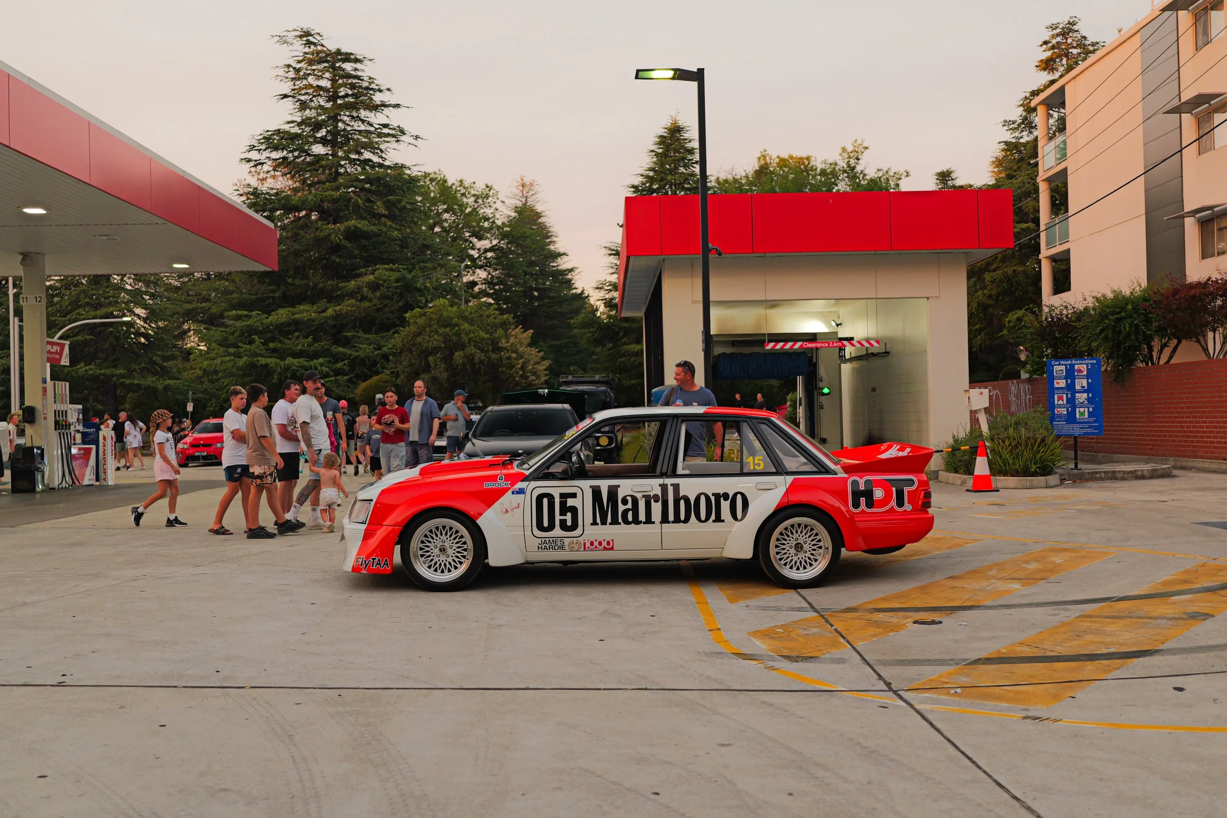 A red and white race car parked in a gas station lot, with people walking around and trees in the background, during dusk.