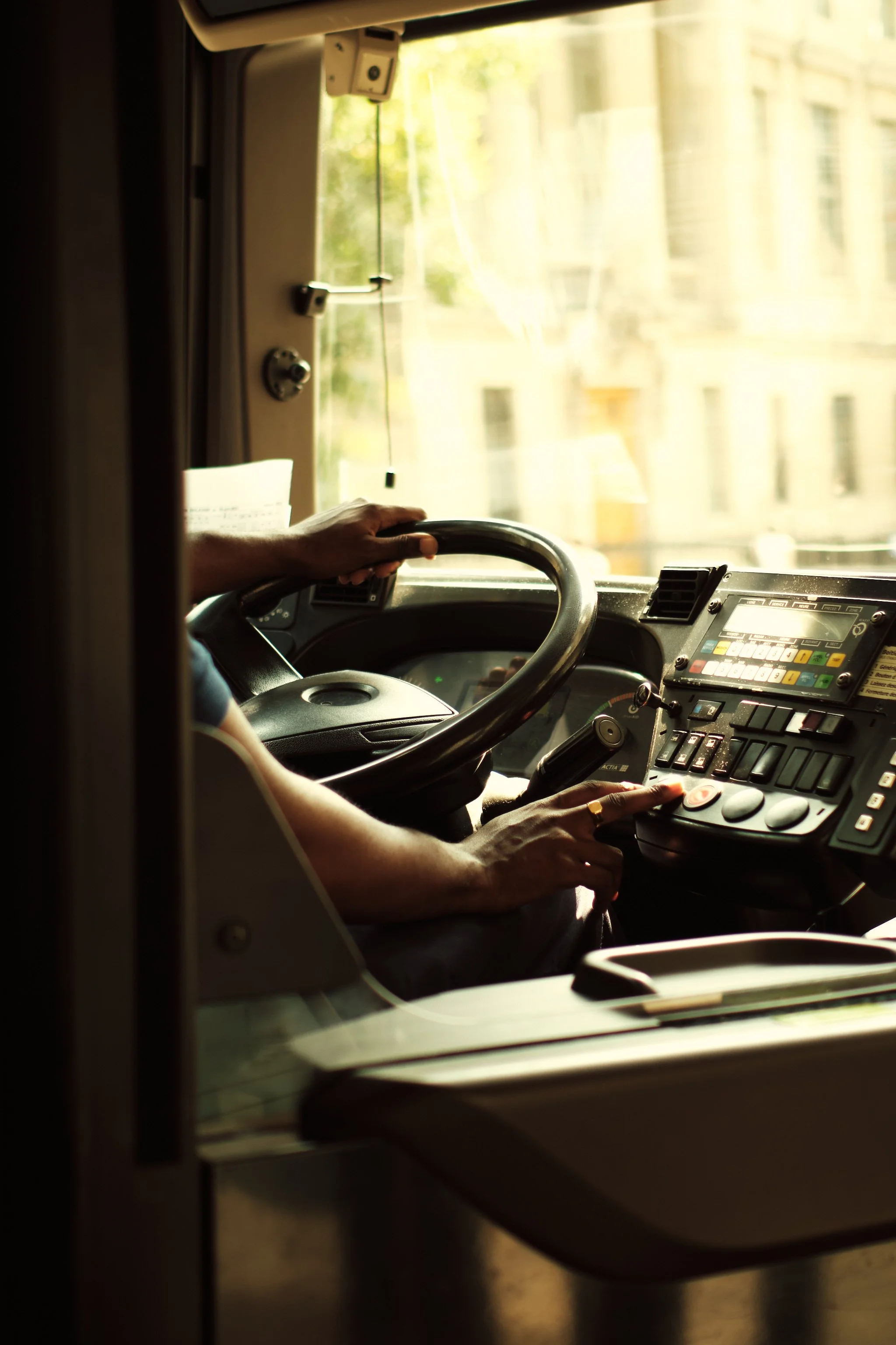 Close-up of a person’s hand on a bus steering wheel, with a control panel and a window showing an urban street scene outside.